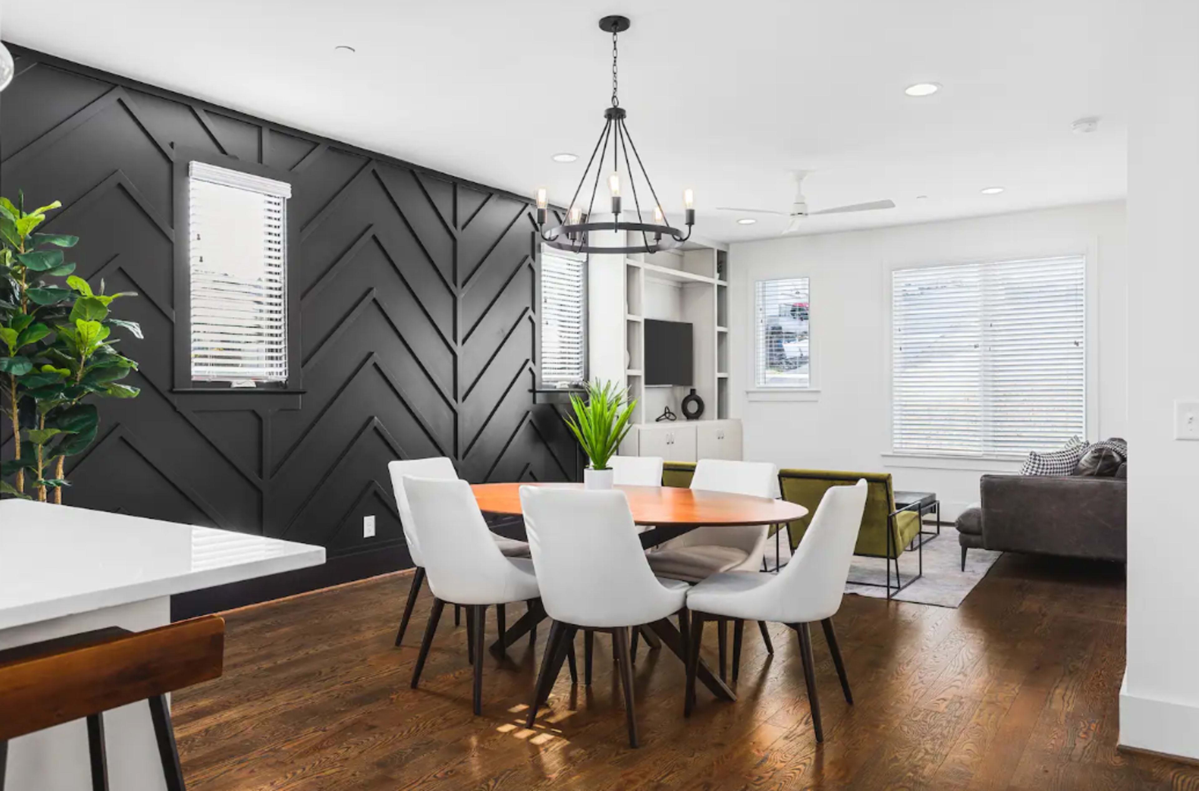 A modern dining area features a round wooden table surrounded by white chairs, with a black accent wall and large windows allowing natural light.