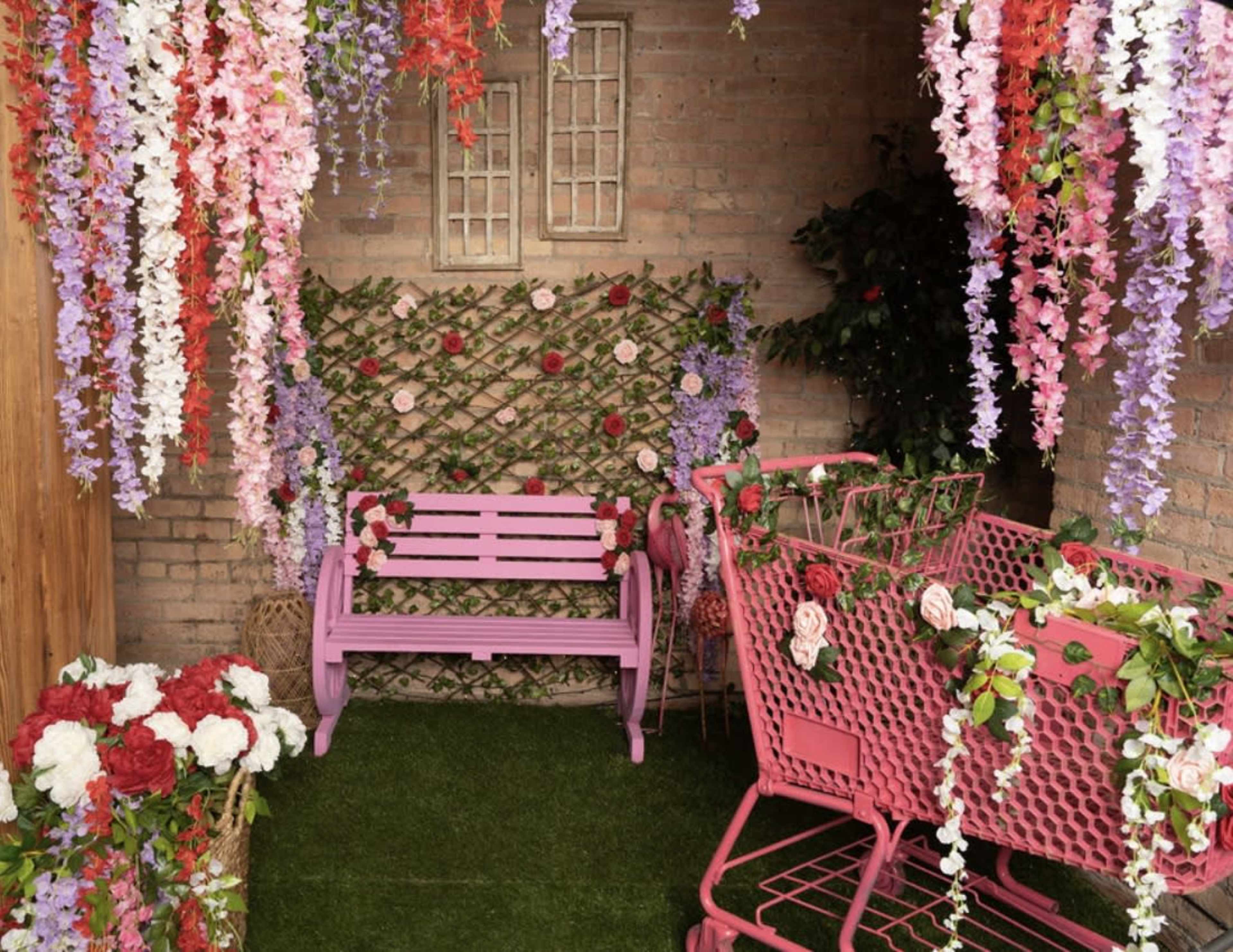 The scene features a decorated corner with a pink bench, two pink shopping carts adorned with flowers, and vibrant hanging floral arrangements against a brick wall.