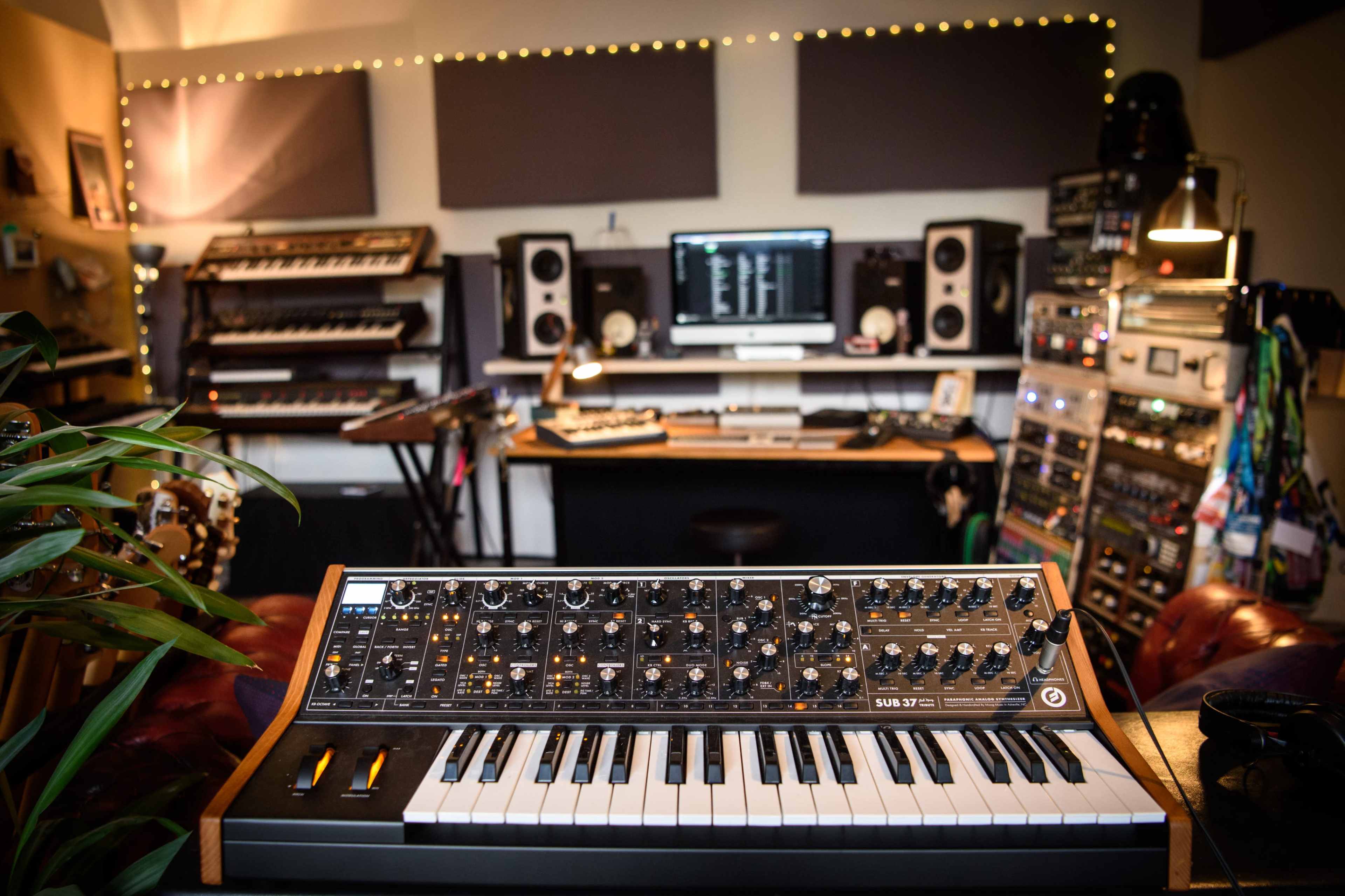 A synthesizer sits in the foreground of a music studio filled with various equipment, monitors, and instruments.