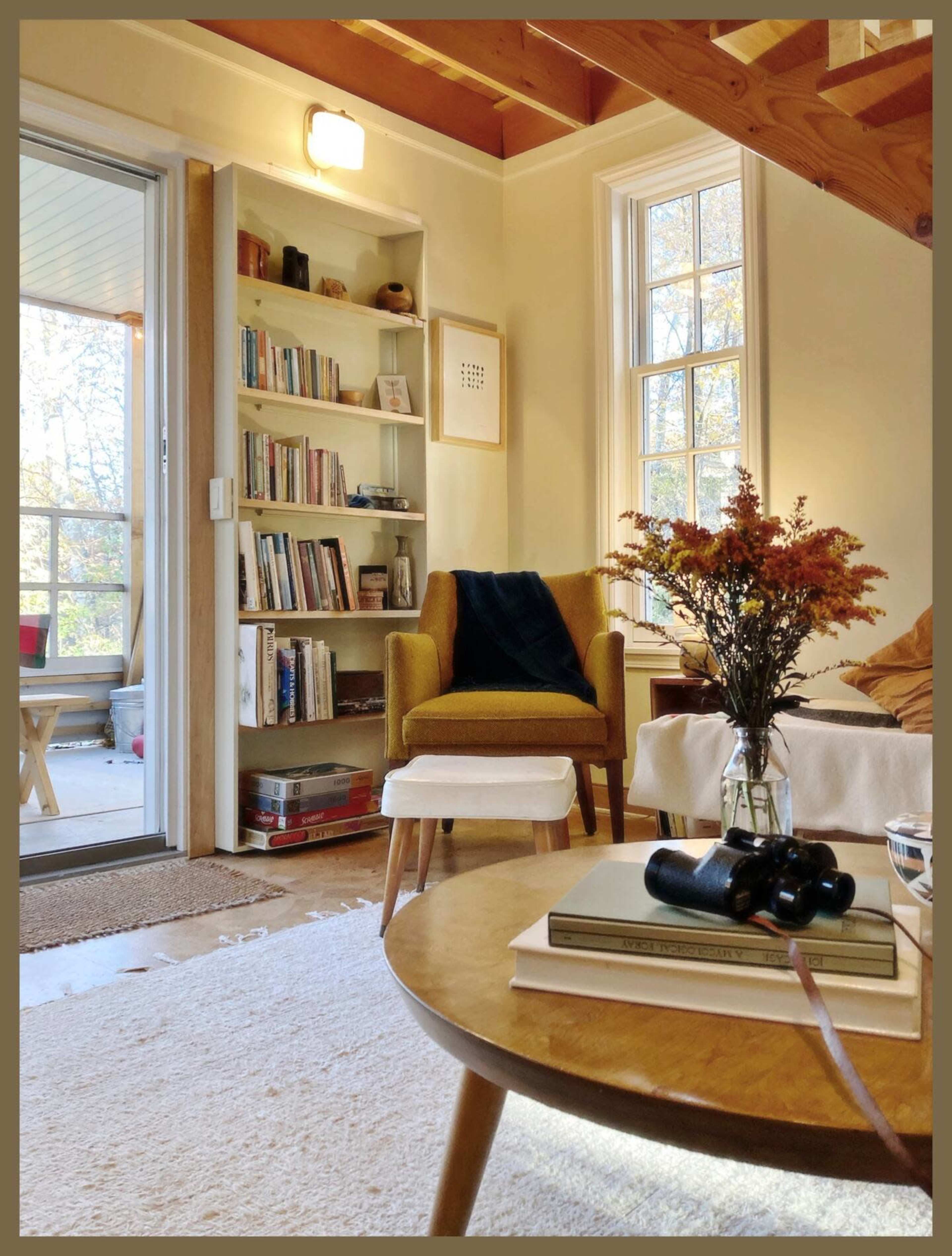 A cozy living room features a bookshelf filled with books, a green armchair, and a coffee table adorned with flowers and stacked books.