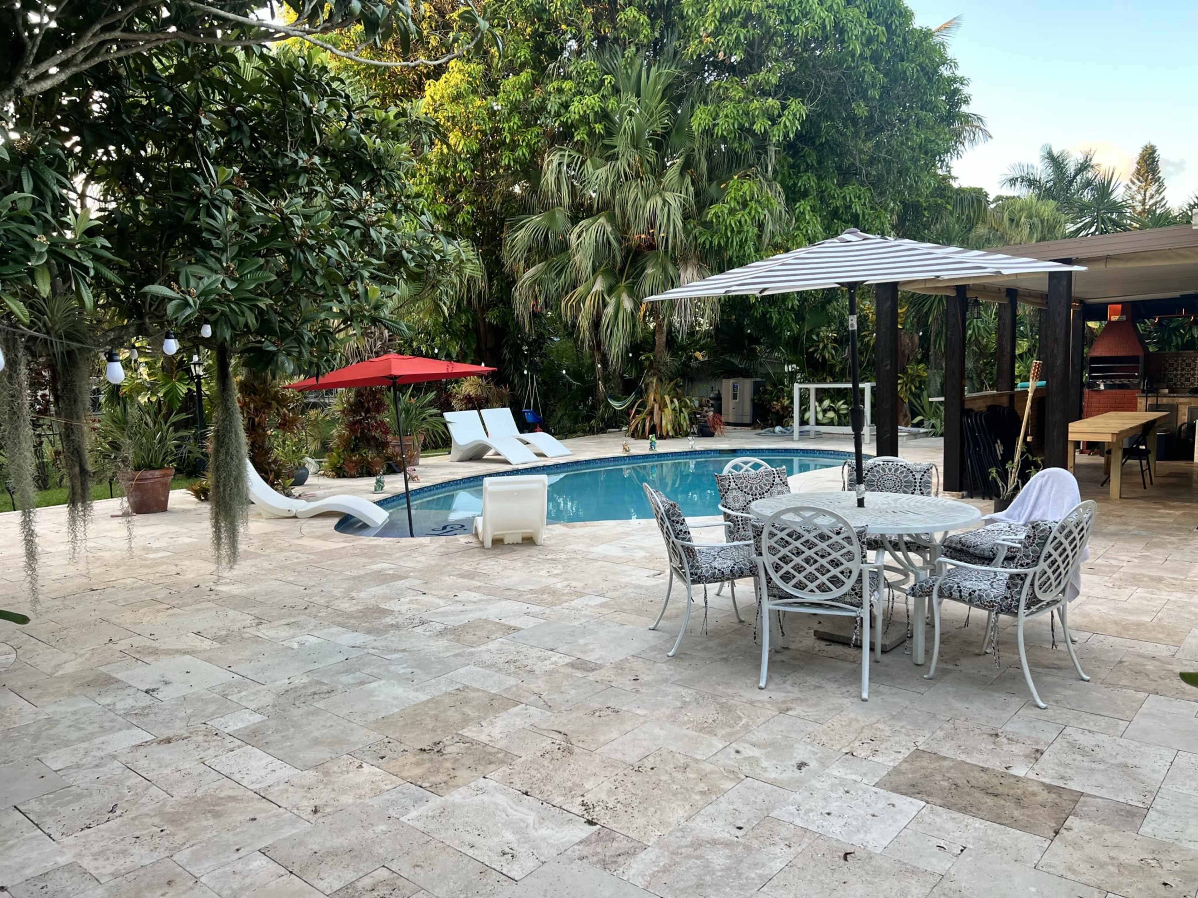 A backyard pool area featuring a circular dining table with chairs, lounge chairs, a red umbrella, and lush greenery surrounding the pool.