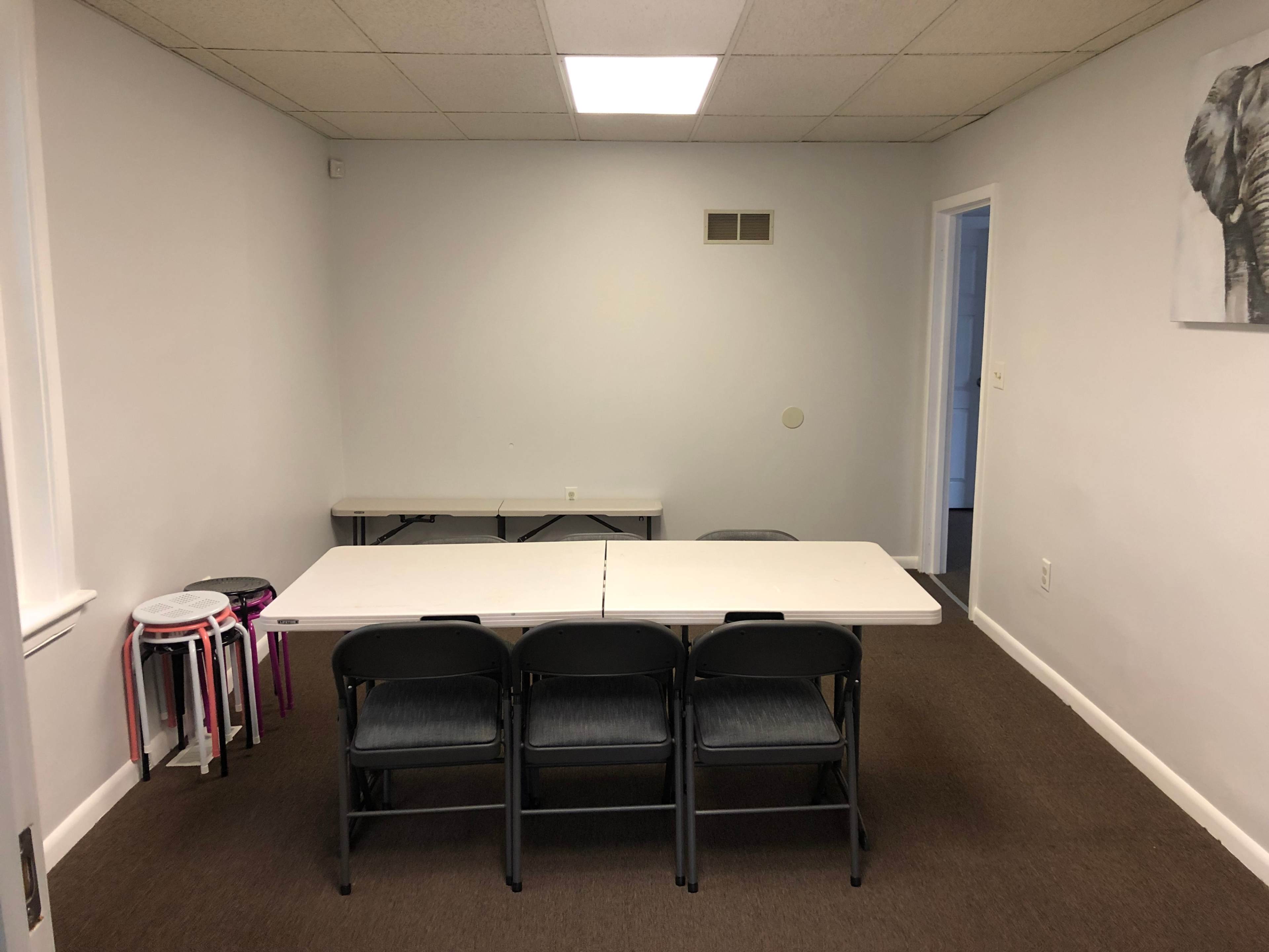 A sparsely furnished room with a long table and four chairs arranged in front of it, alongside a few stools and an elephant artwork on the wall.