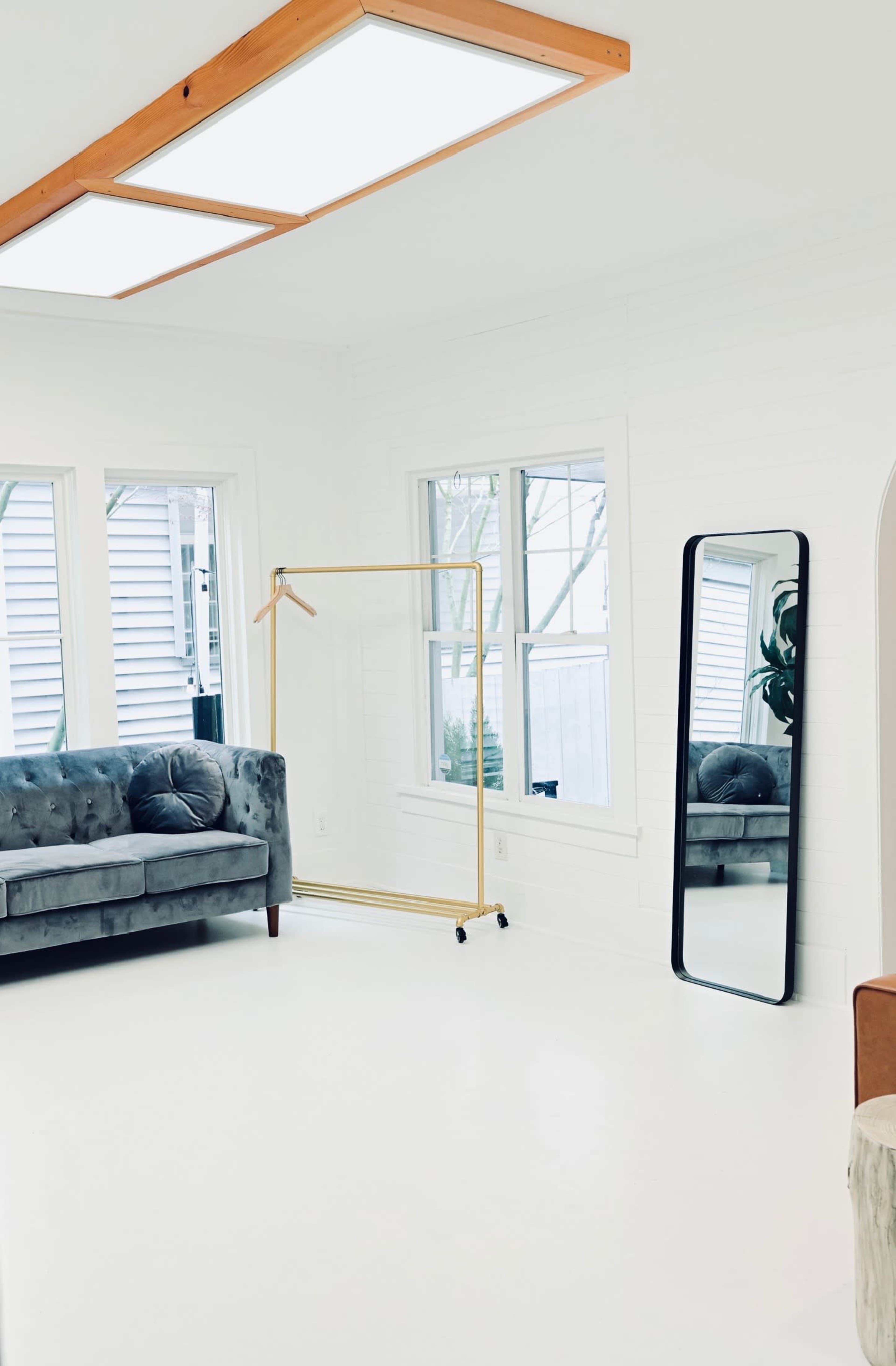 The image shows a bright, minimalistic room with a gray velvet sofa, a gold clothing rack on wheels, and a tall mirror against a white wall.