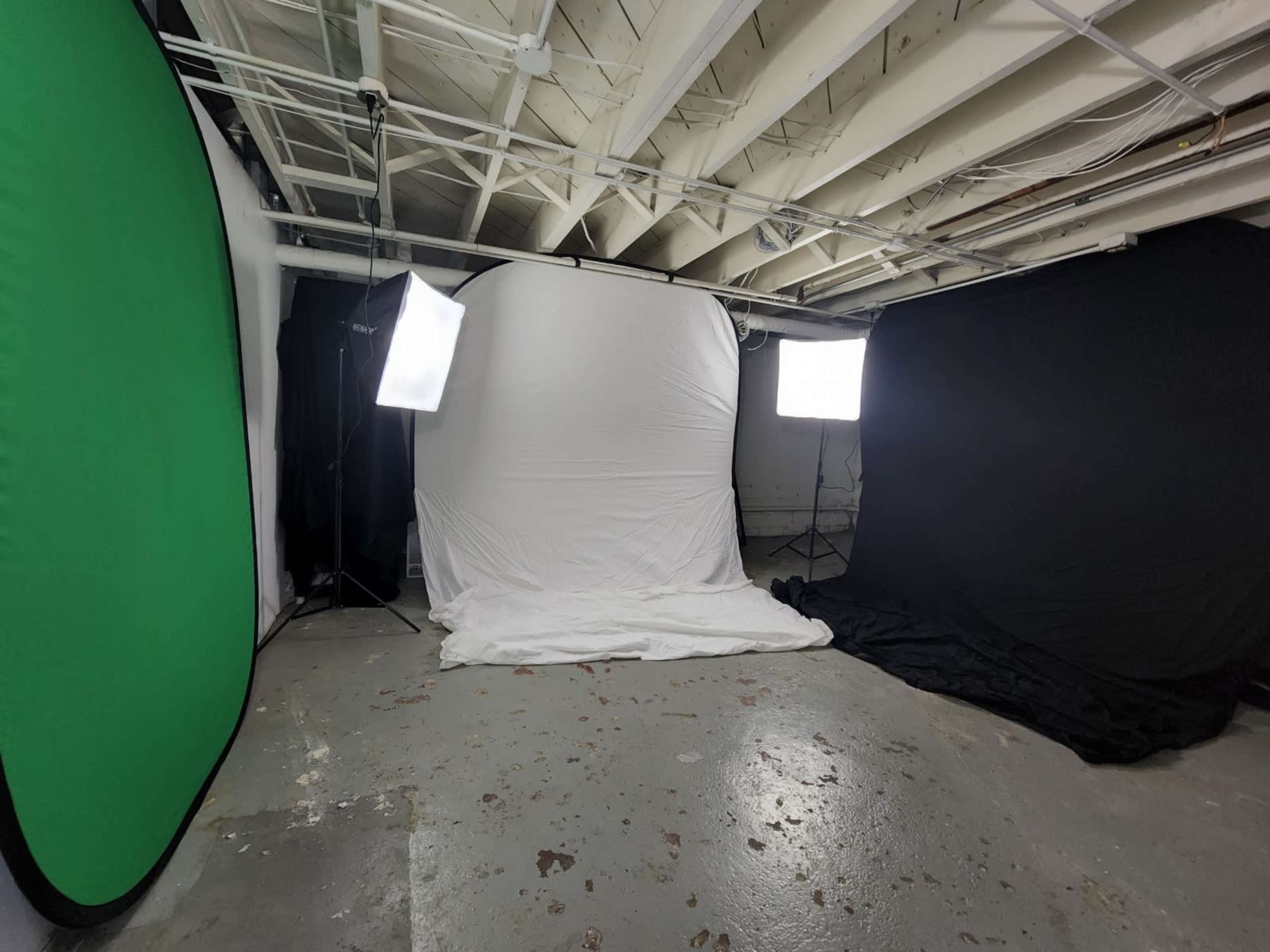 A photography setup in a basement, featuring a white backdrop surrounded by a green backdrop, a black backdrop, and two softbox lights.