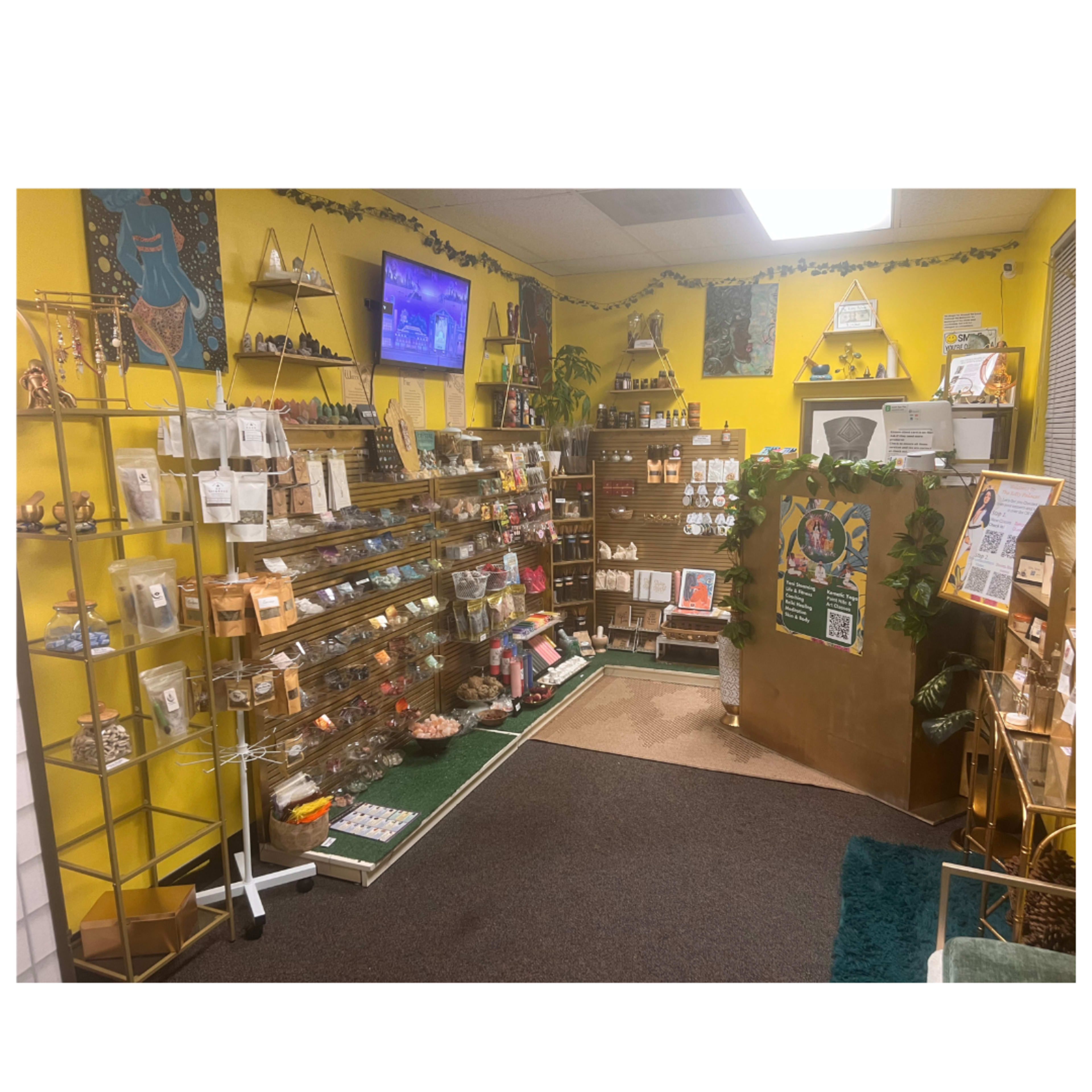 The image shows a brightly colored retail space filled with shelves stocked with various products, including crystals, herbs, and wellness items, alongside plants and a television mounted on the wall.