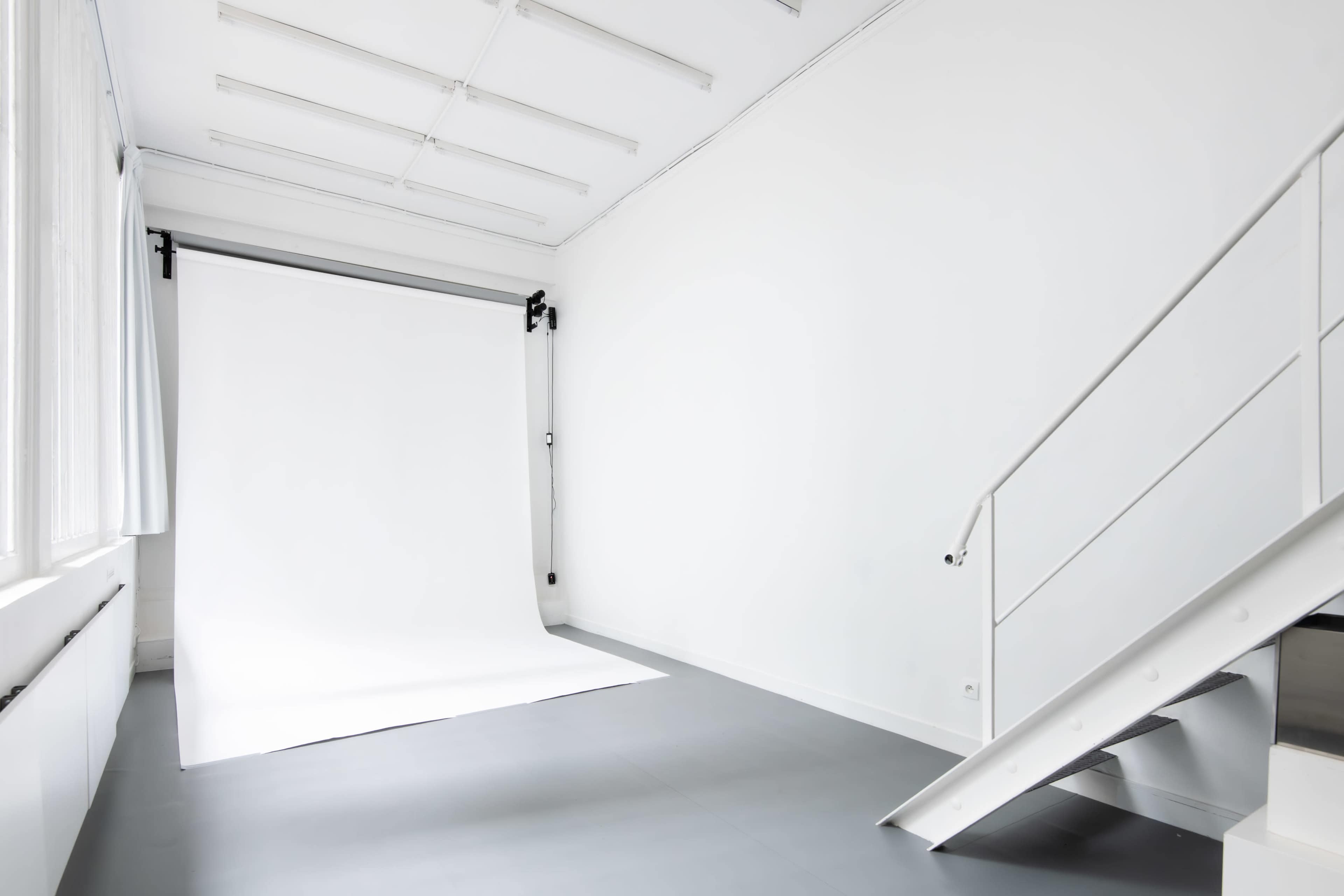 The image shows a minimalist white studio space with a backdrop and a metal staircase.