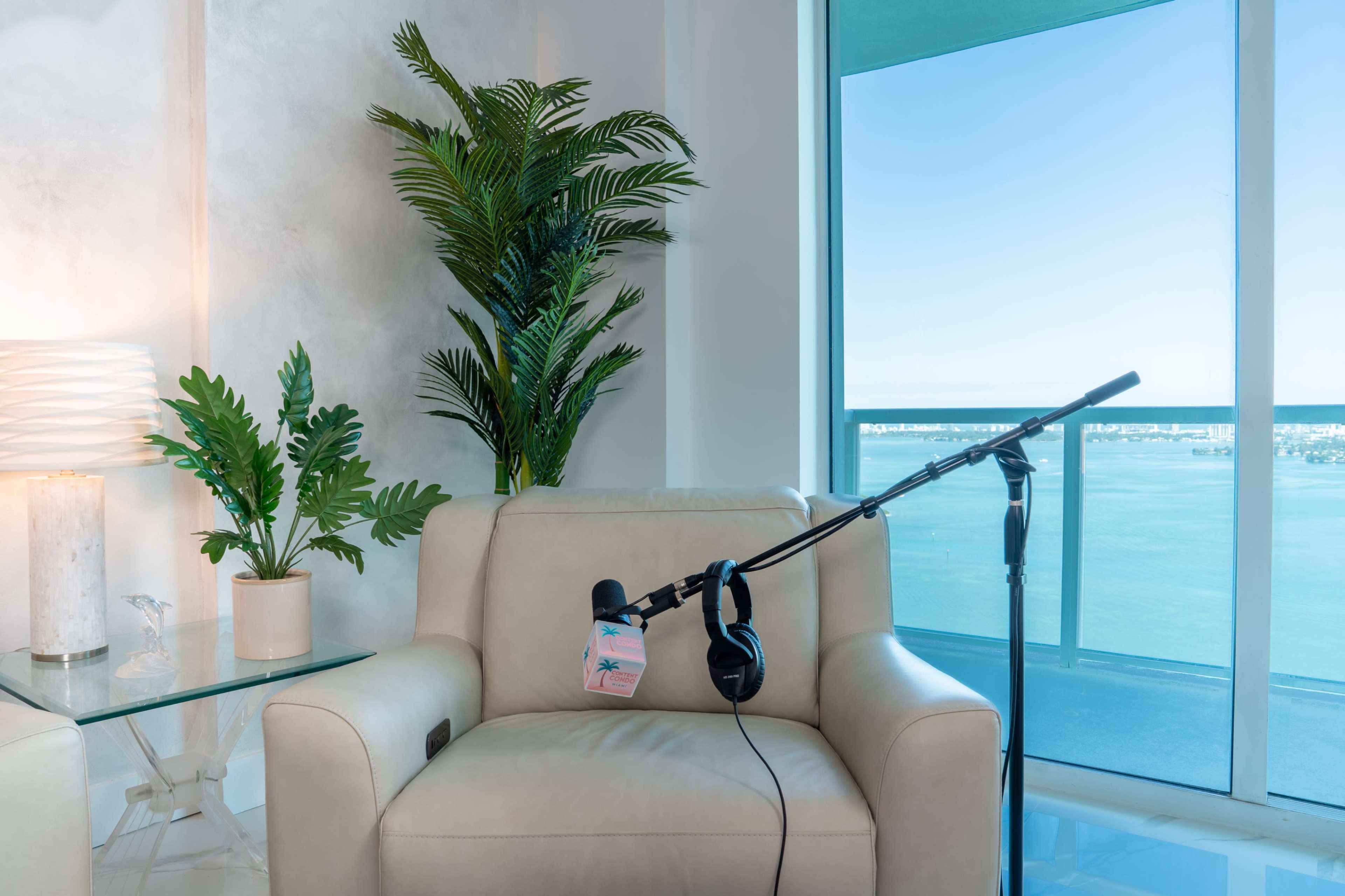 A microphone and headphones are resting on a chair next to a glass table with plants, against a backdrop of large windows overlooking a body of water.
