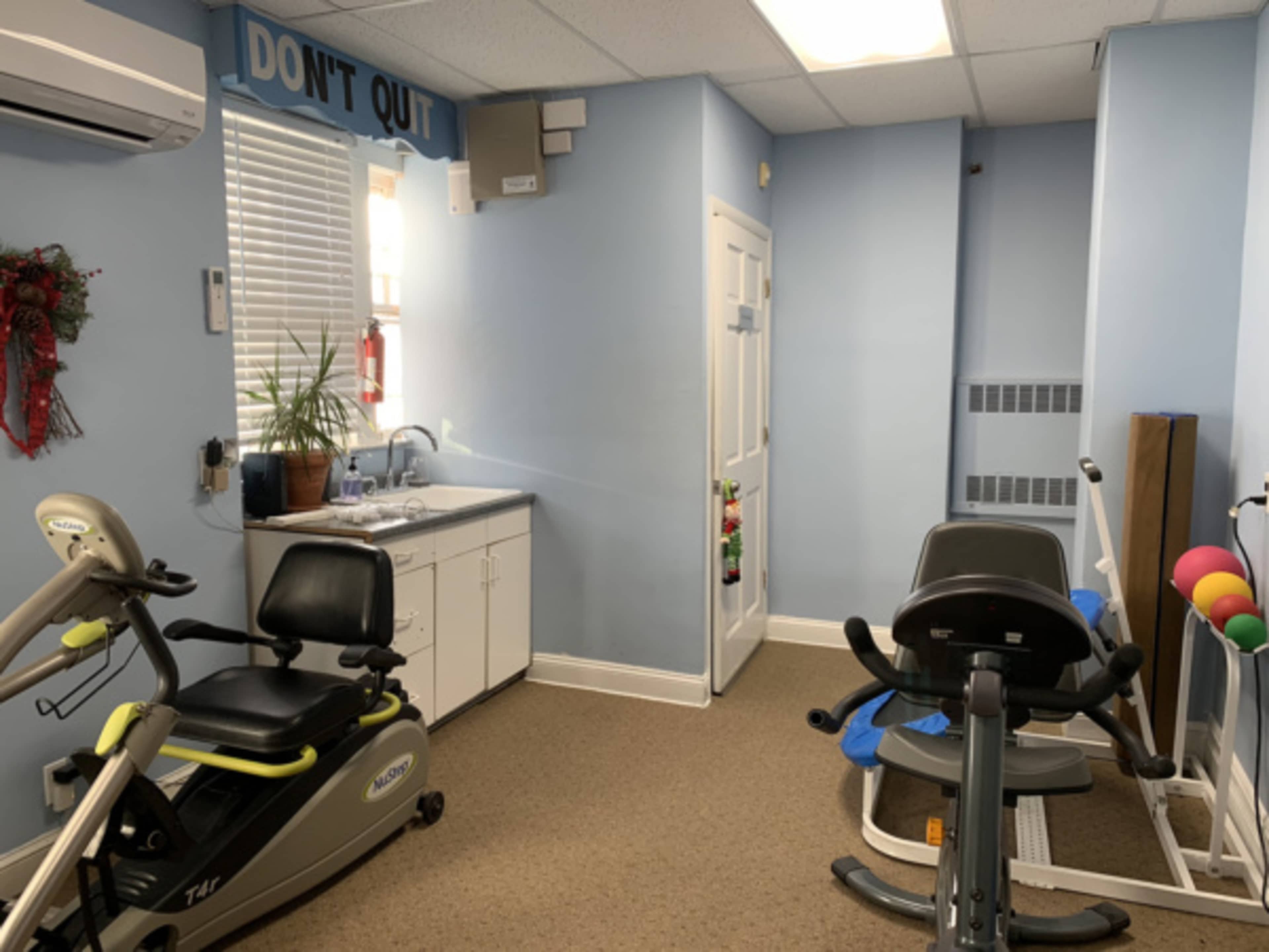 The image shows a small, light blue exercise room equipped with fitness machines and a sink, featuring a sign that reads "DON'T QUIT" on the wall.