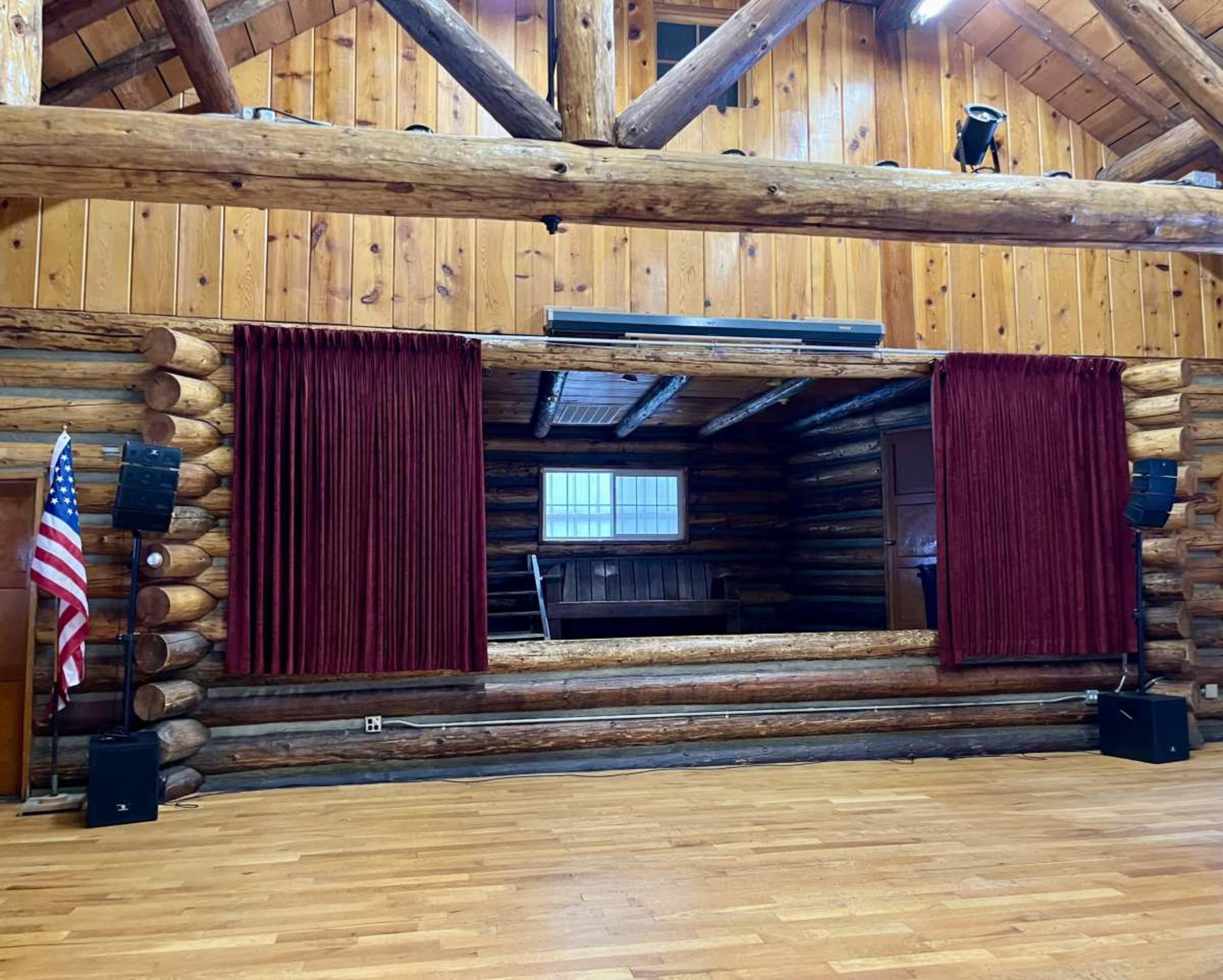 The image shows a wooden interior of a performance space with a stage framed by red curtains, flanked by sound equipment and an American flag on one side.