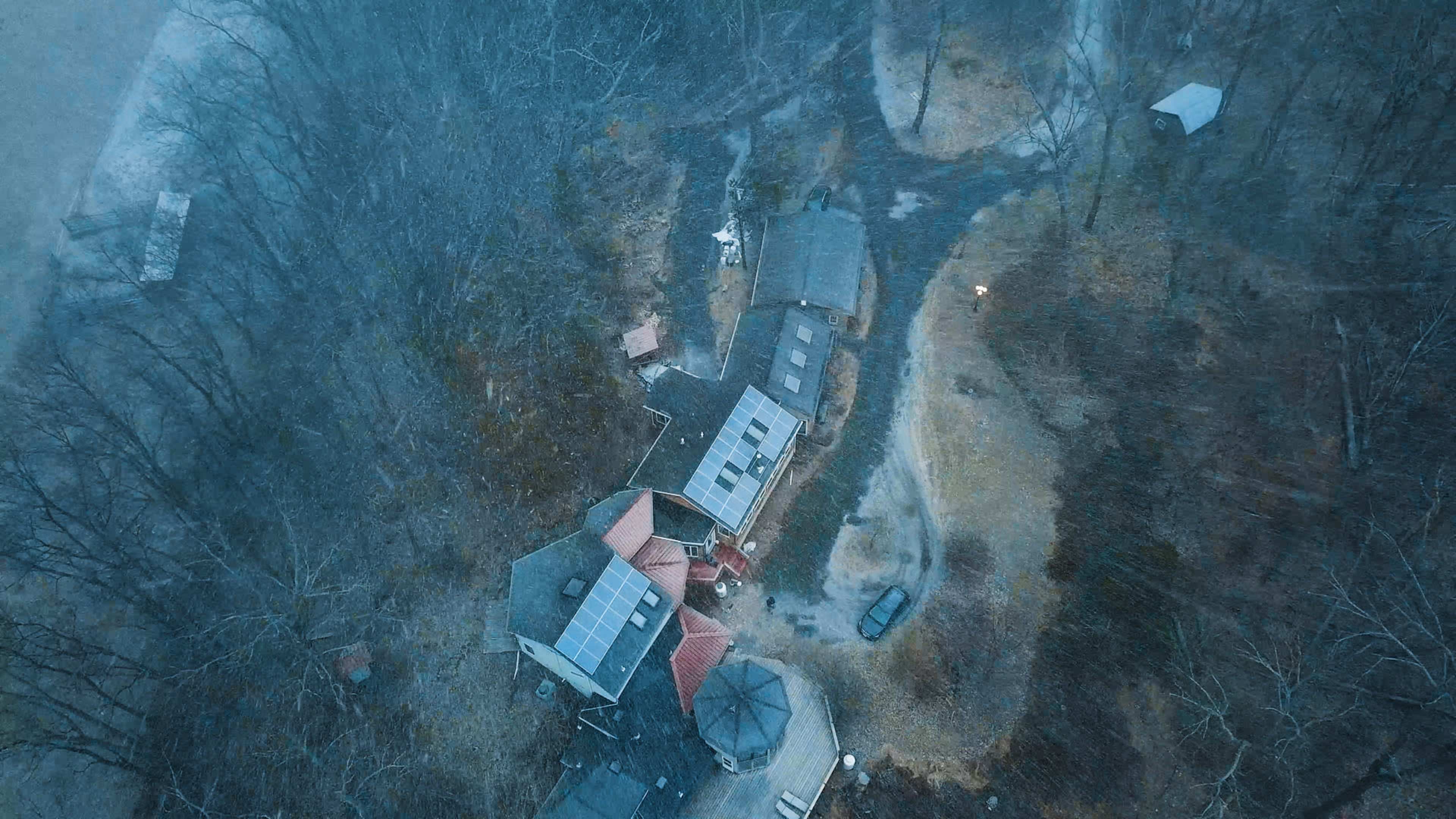 An aerial view shows several buildings, including solar-paneled roofs, surrounded by bare trees and a winding path in a rural area.