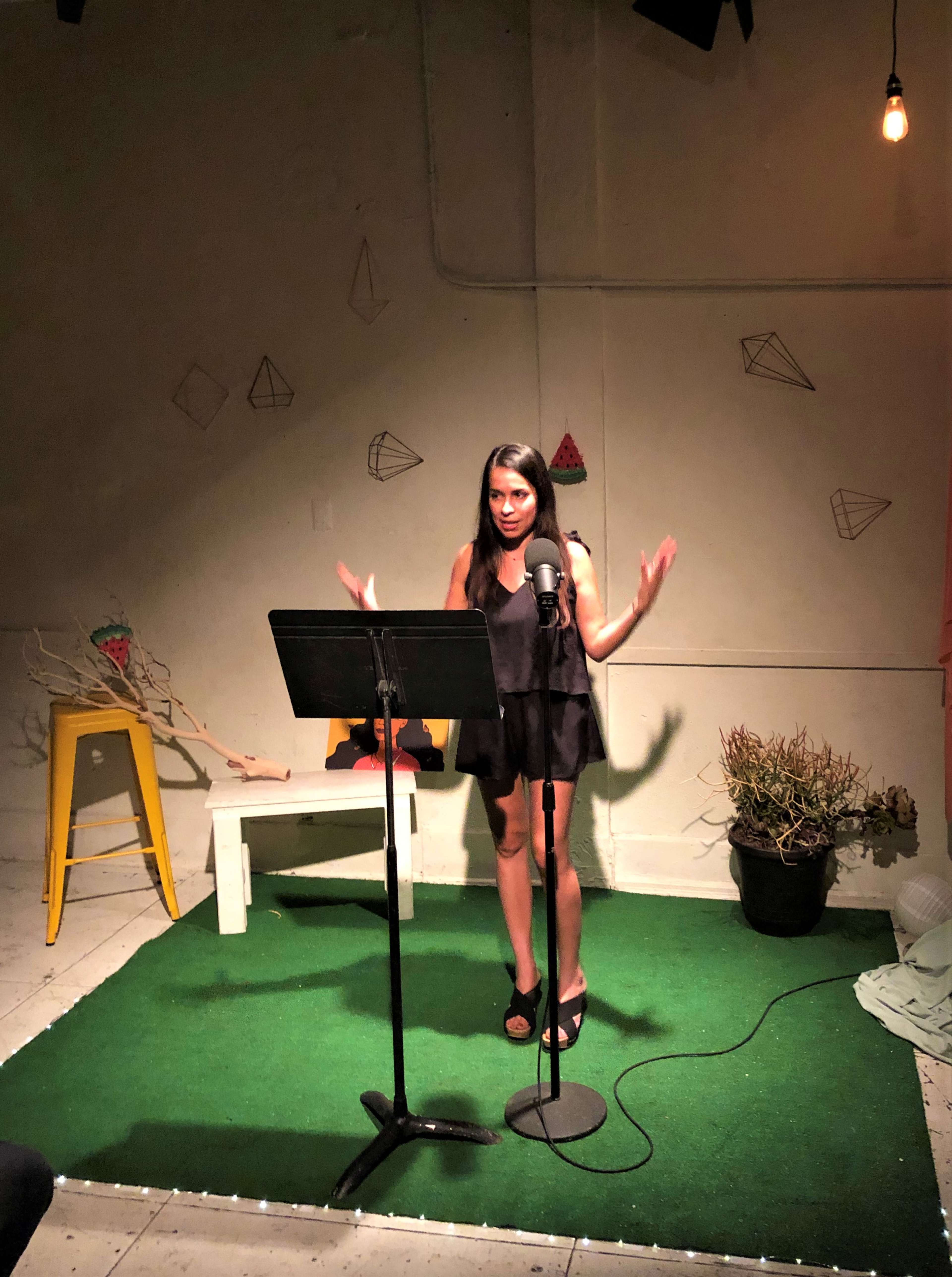 A woman stands on a small stage with a microphone, gesturing as she speaks in a room decorated with geometric shapes.