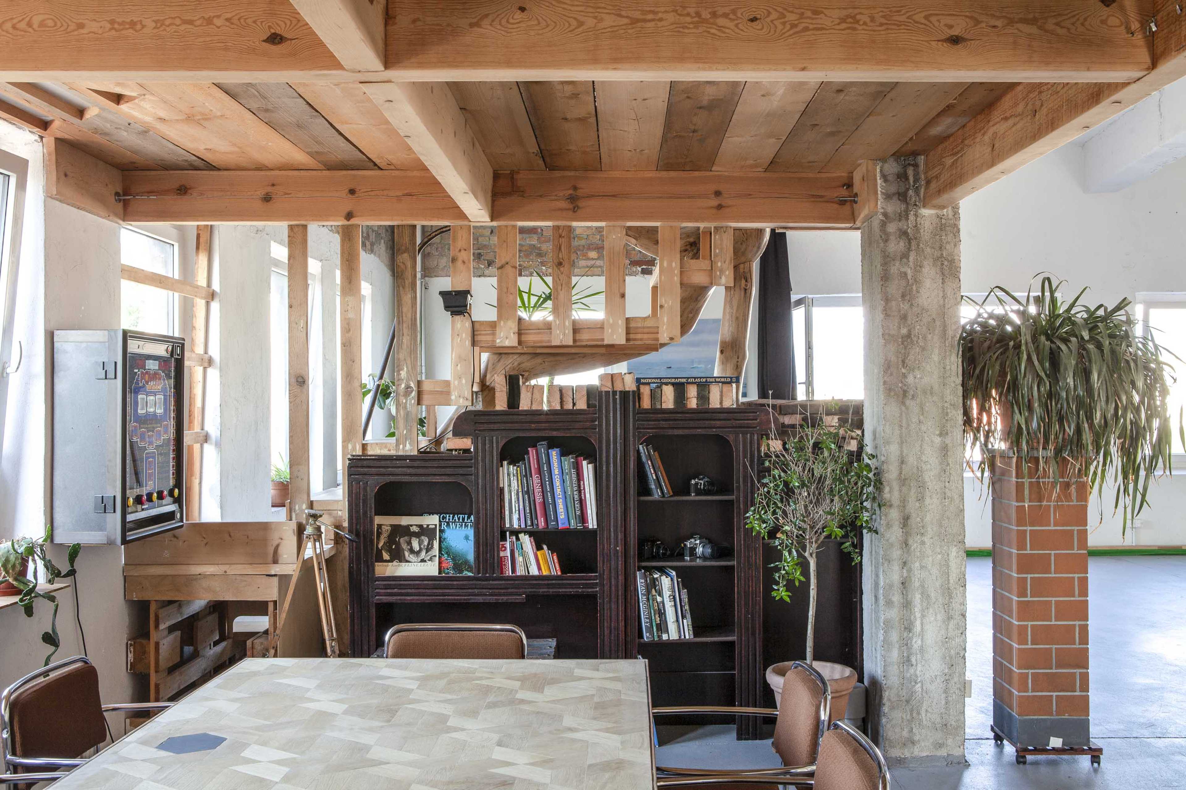 An open, modern interior space featuring wooden beams, a table with a geometric pattern, a bookshelf filled with books, and large windows.