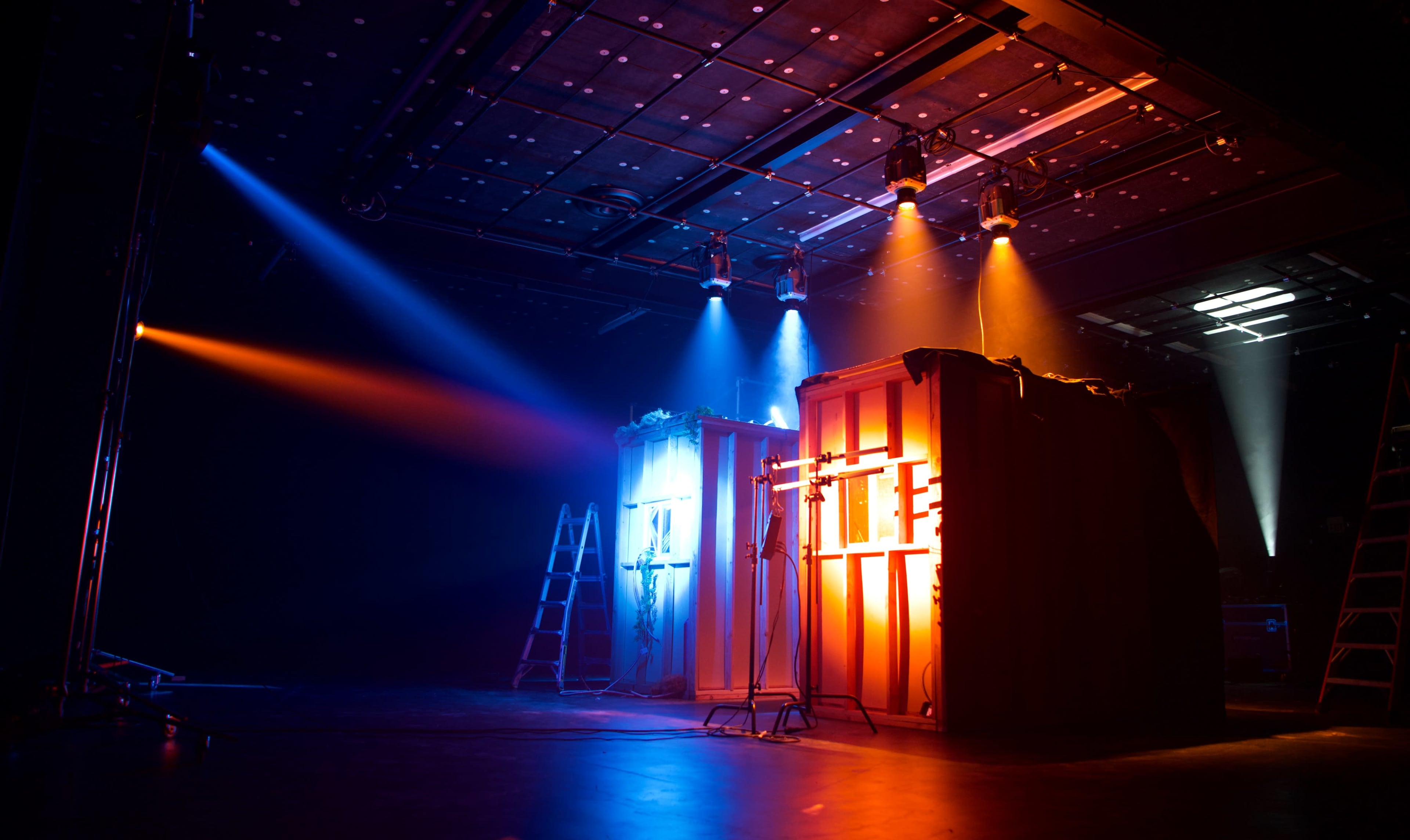 Two lighted, wooden structures sit on stage, illuminated by blue and orange spotlights, with ladders positioned nearby.