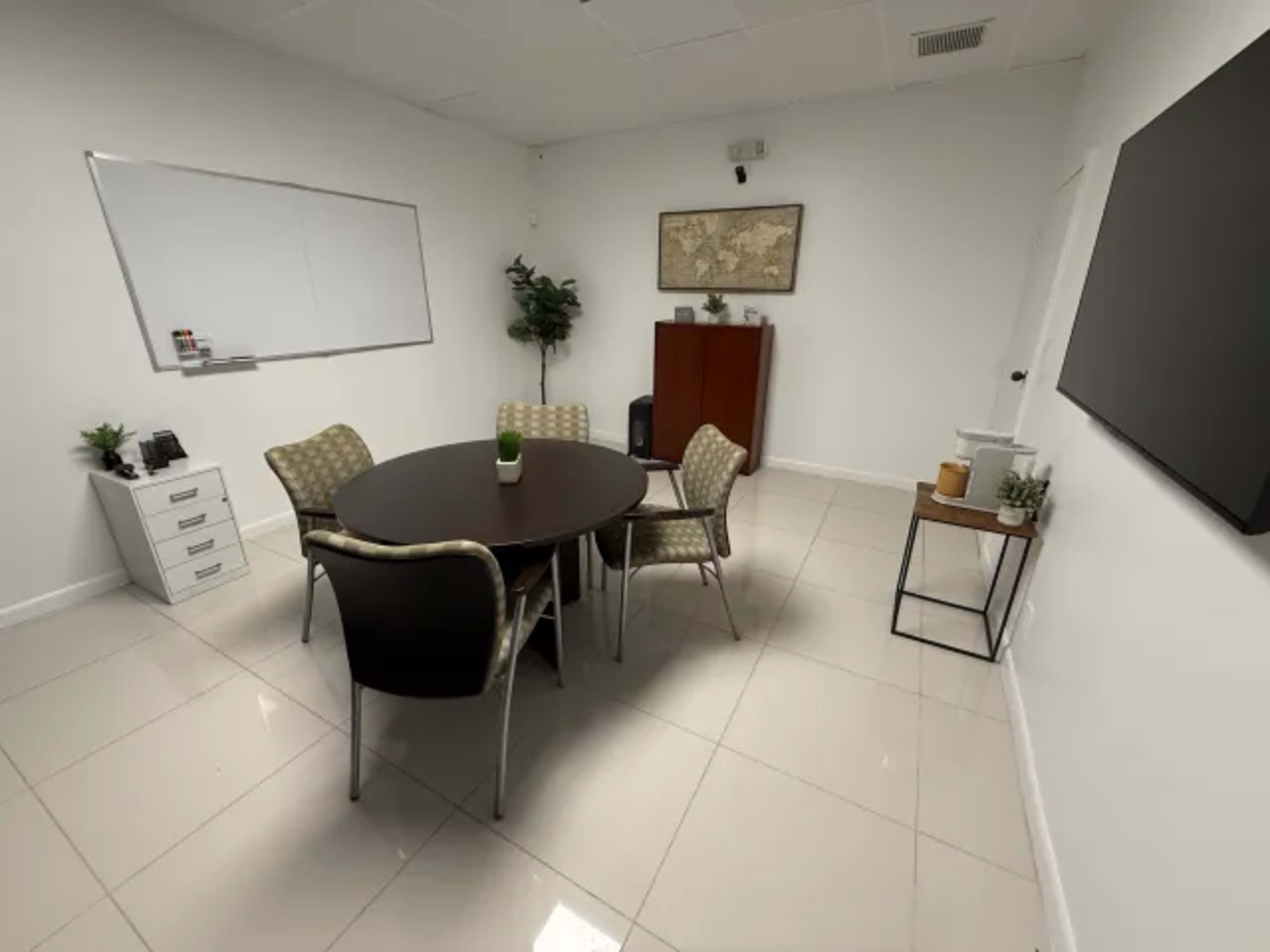 The image shows a modern conference room featuring a round table with four chairs, a whiteboard on the wall, a cabinet, and several indoor plants.