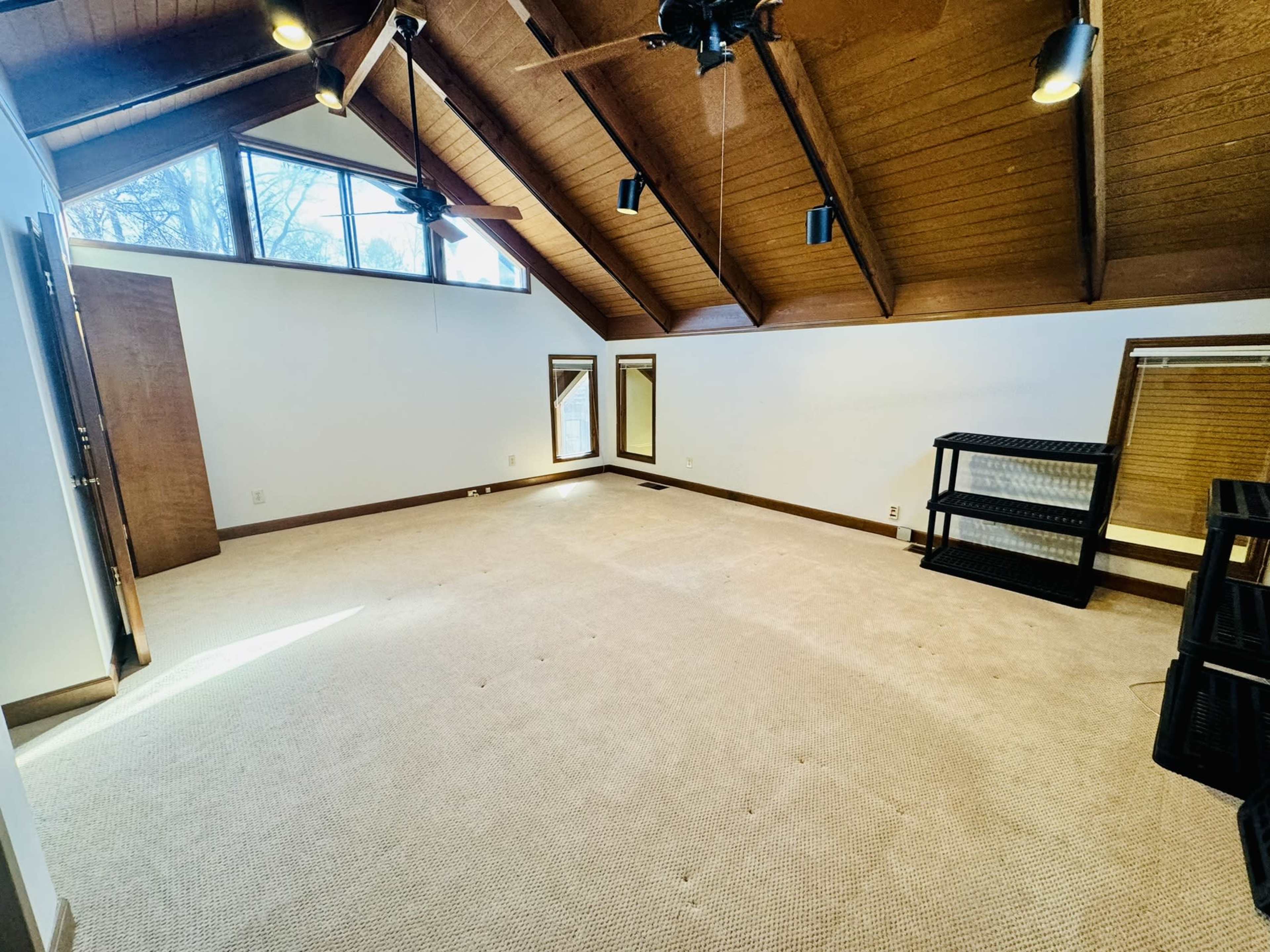 The room features a vaulted wooden ceiling, large windows providing natural light, and a carpeted floor with minimal furniture.