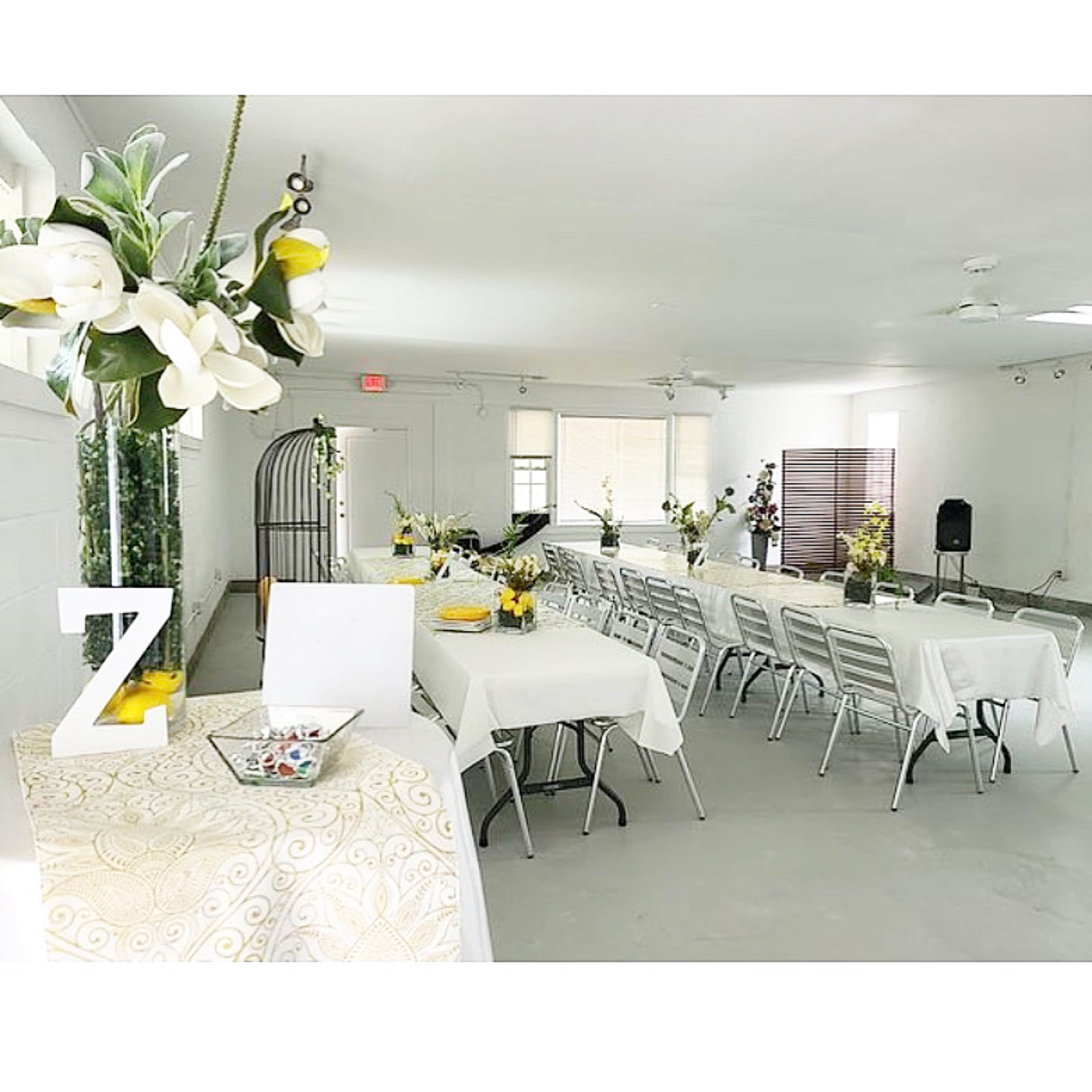 A bright event space features long tables set with white linens and floral centerpieces, with a small decorative table in the foreground.