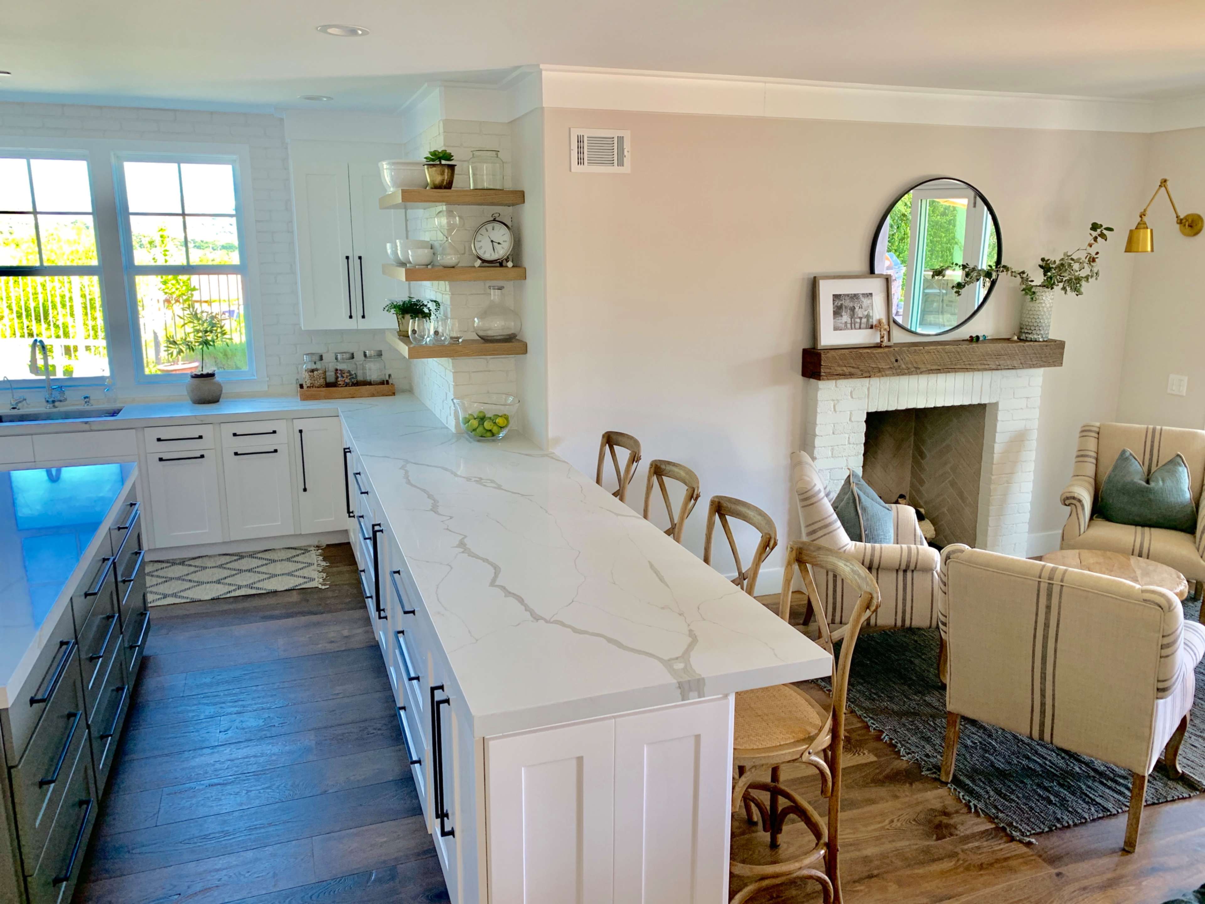 A modern kitchen featuring a large island with a white countertop, a cozy seating area with chairs beside a fireplace, and a window providing natural light.