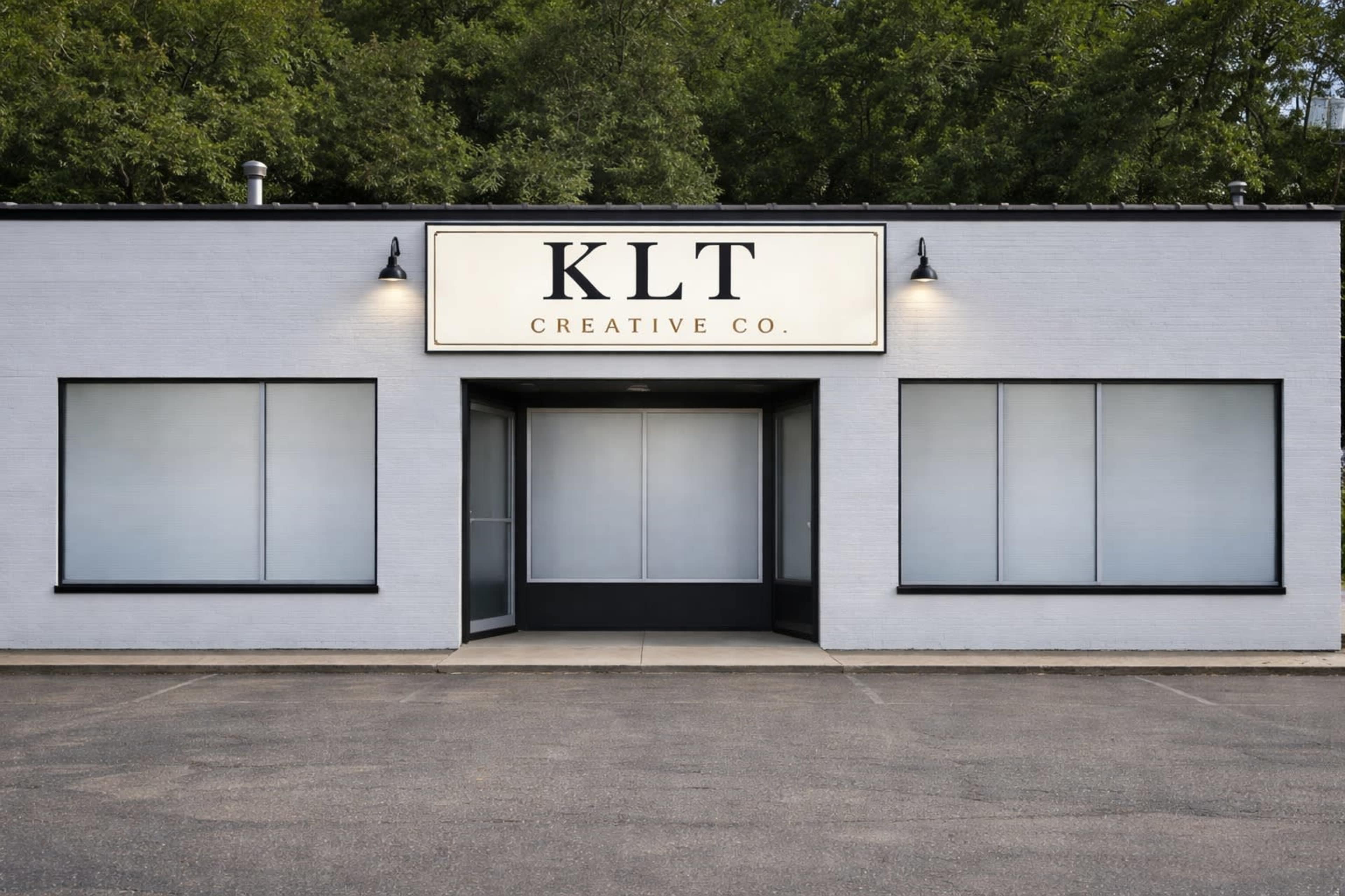 The image shows a single-story building with a sign that reads "KLT CREATIVE CO" above the entrance, flanked by large, blank windows and surrounded by greenery.