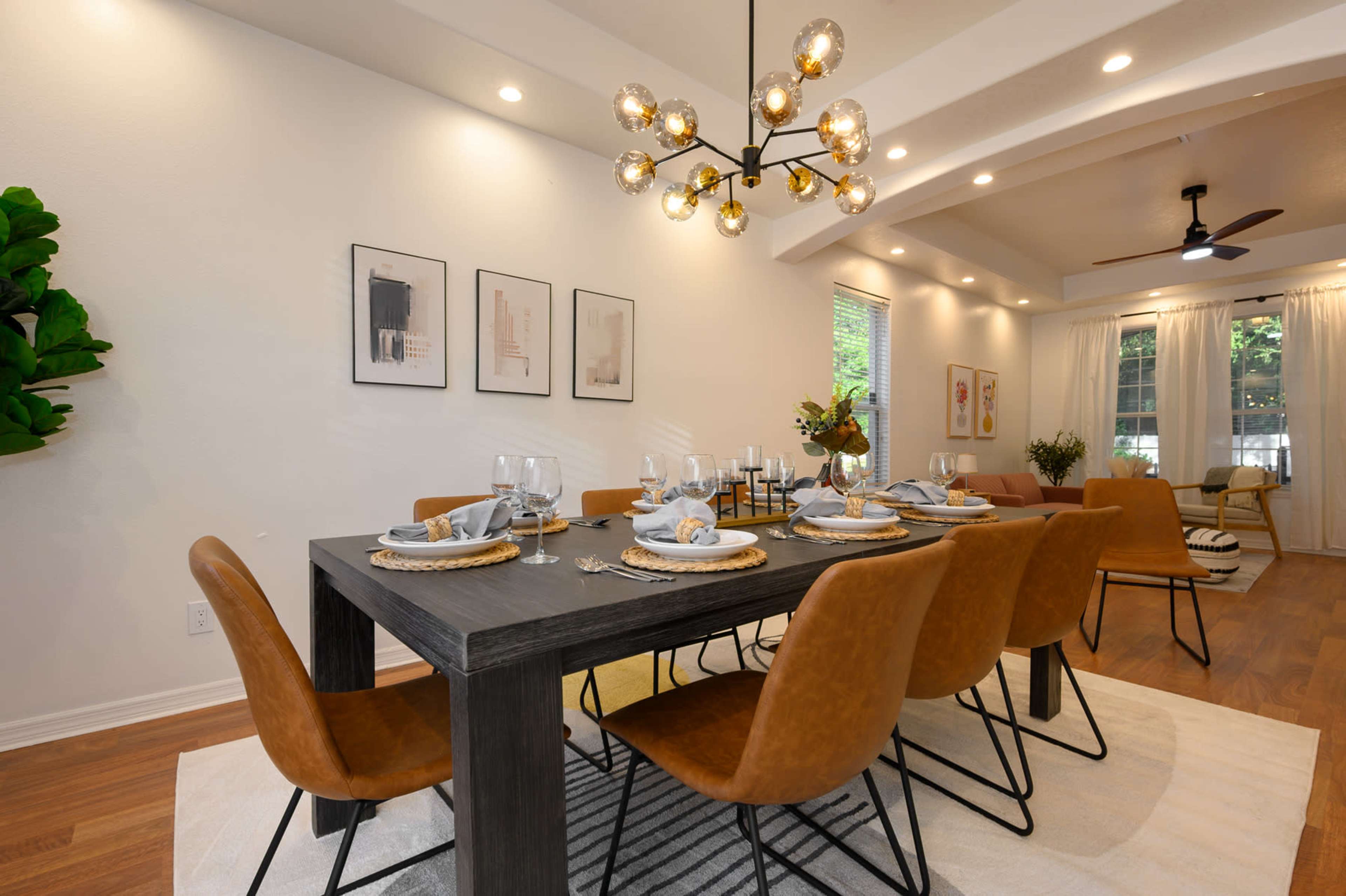 A modern dining room features a large table set with dinnerware and surrounded by stylish brown chairs, with a decorative light fixture above and artwork on the walls.