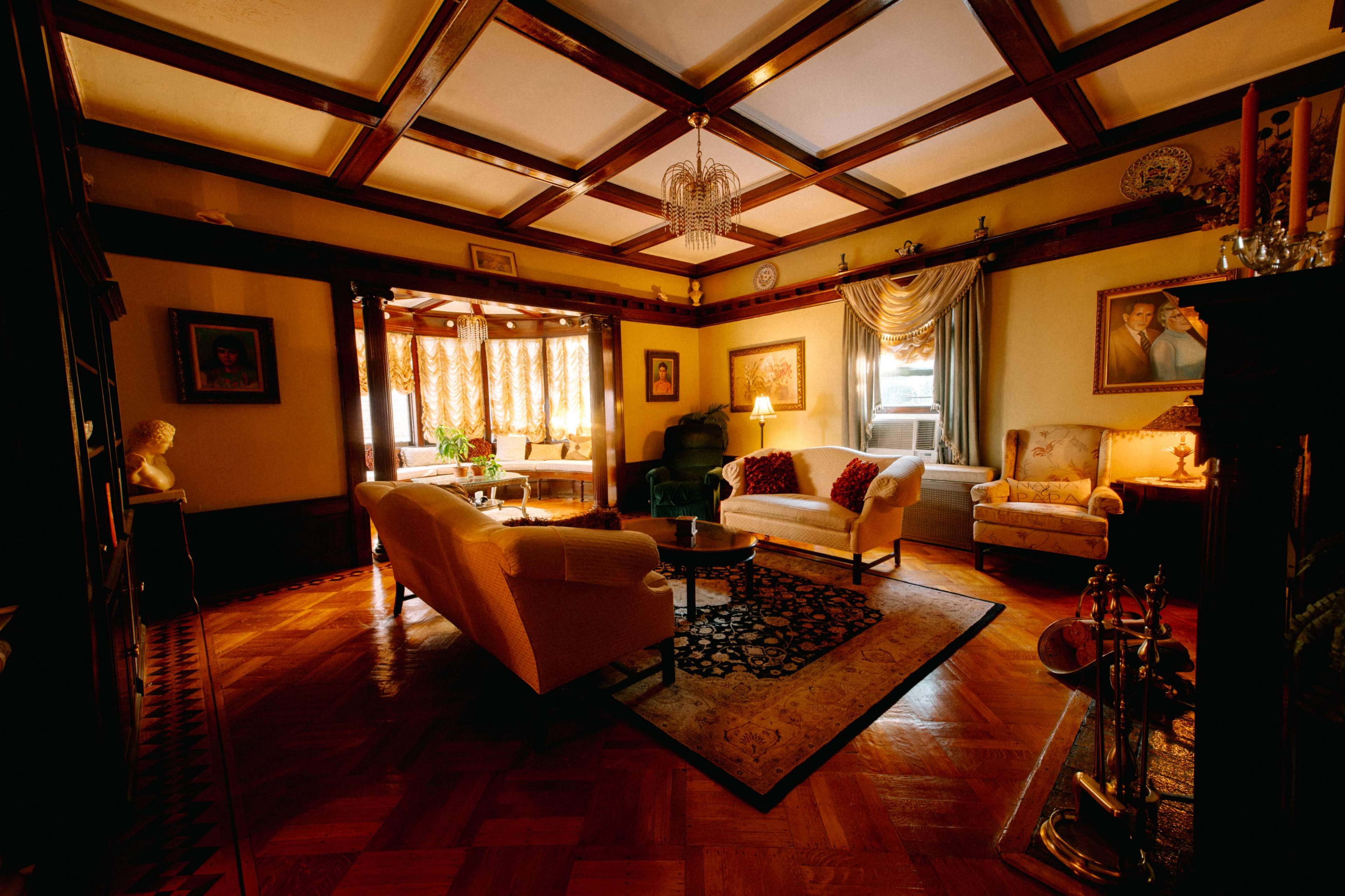 The image shows a richly decorated living room featuring a coffered ceiling, comfortable seating, and warm lighting.