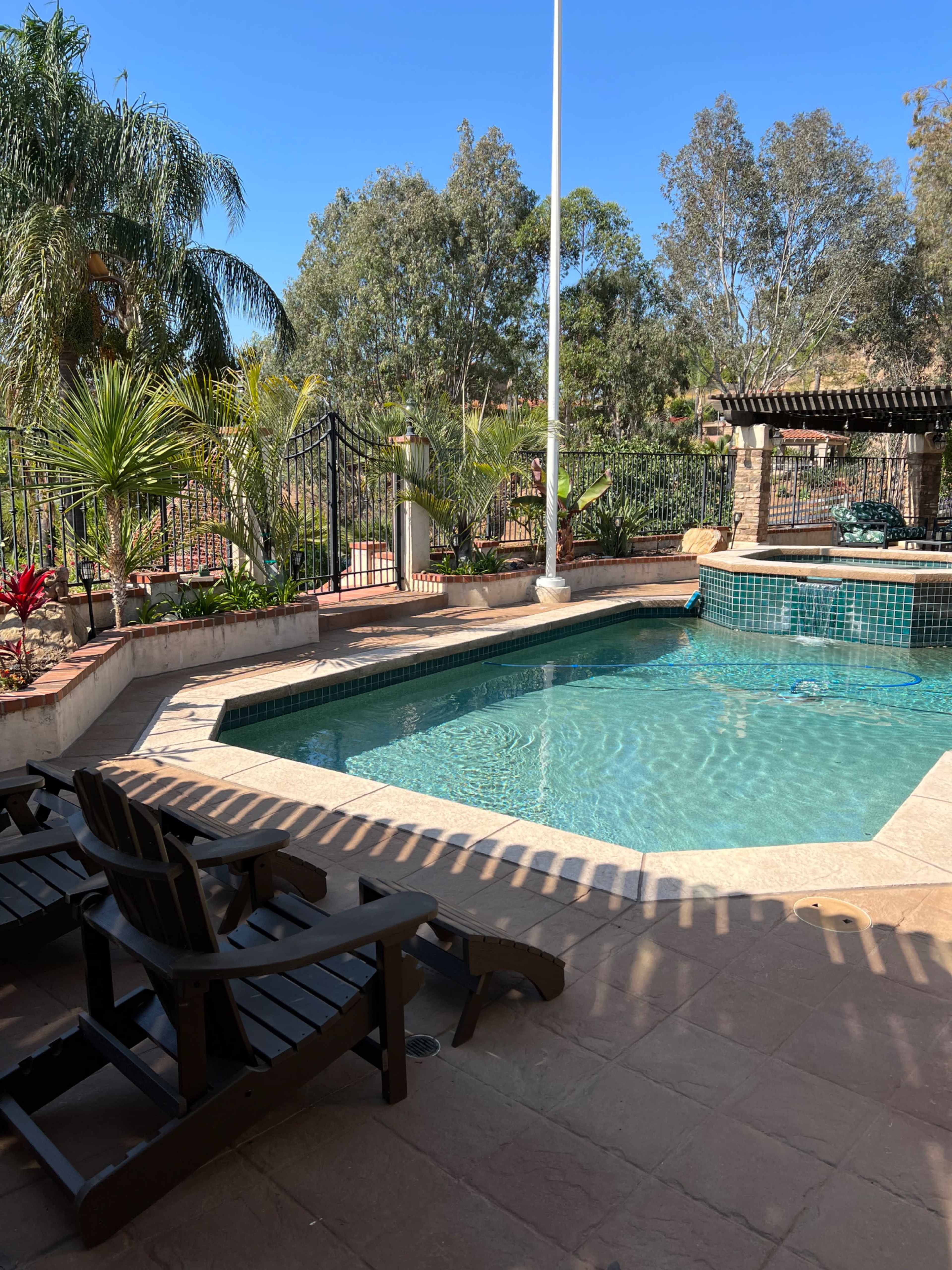 The image shows a residential pool area surrounded by lush greenery and palm trees, with two wooden lounge chairs positioned nearby.