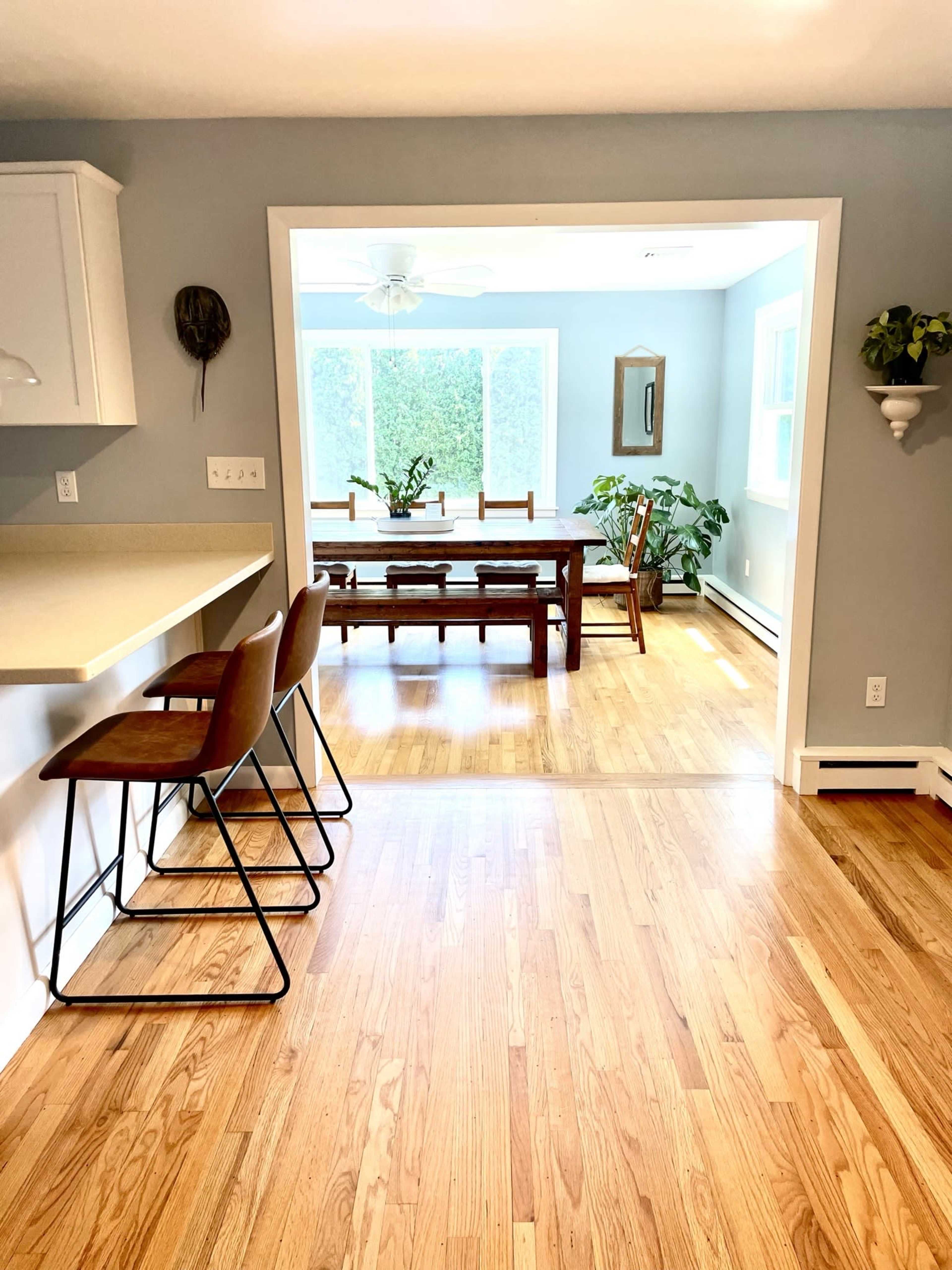 A kitchen with bar stools overlooks a dining area featuring a large wooden table and plants, separated by a doorway.