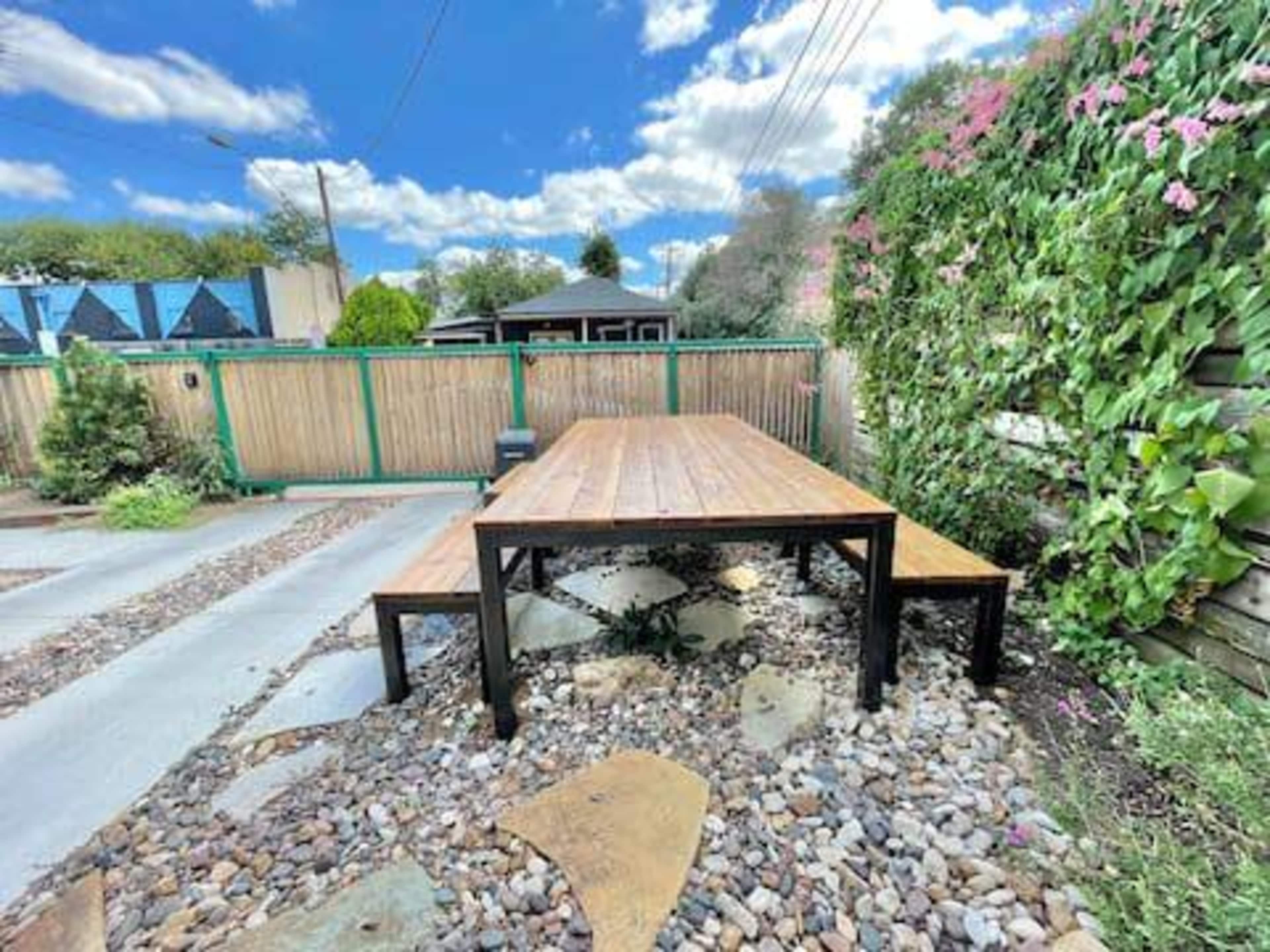 A wooden picnic table with benches is set on a gravel surface surrounded by greenery and a wooden fence.