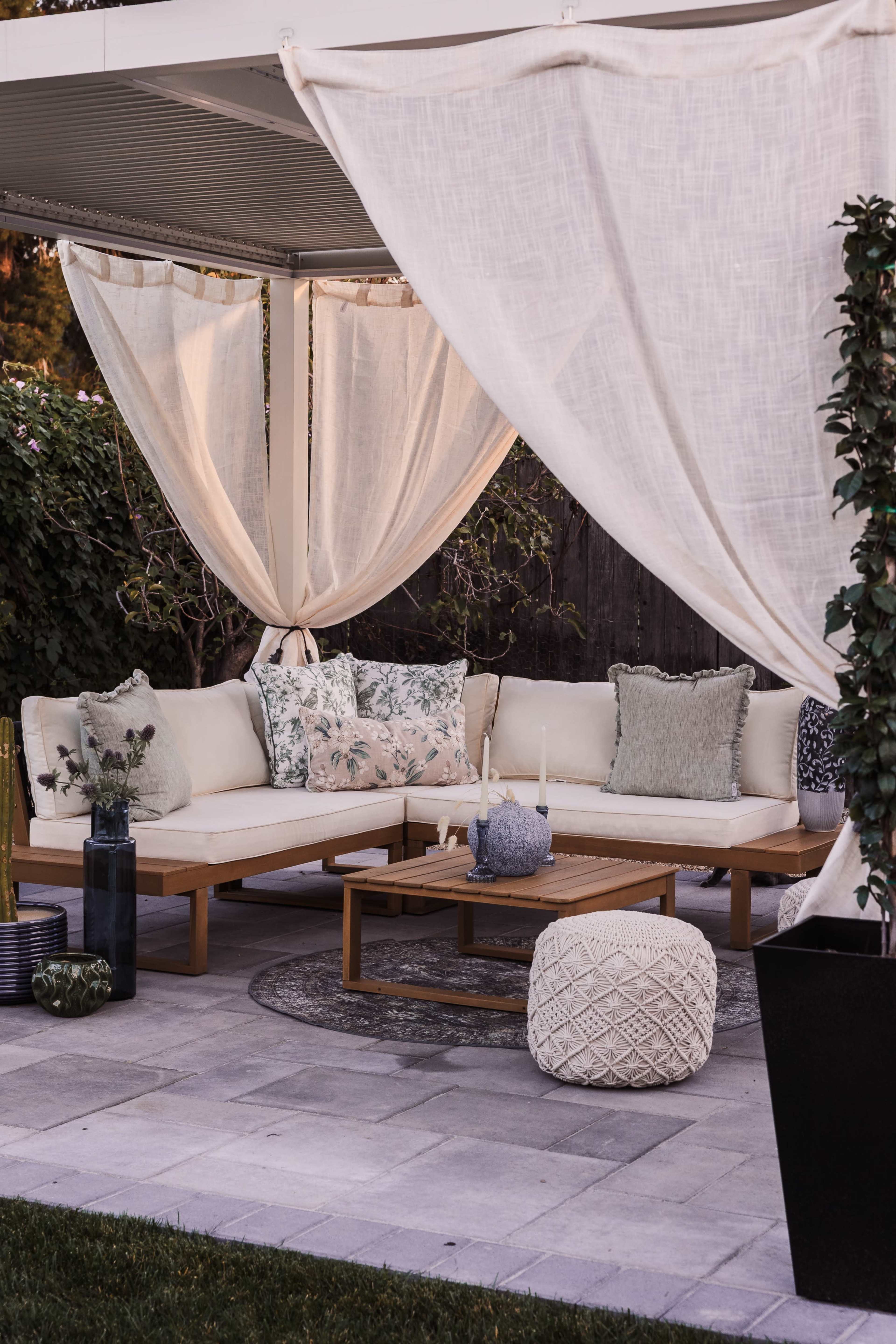 The image shows a cozy outdoor seating area with a white fabric canopy, featuring cushioned sofas, a wooden coffee table, and decorative pillows, surrounded by greenery.