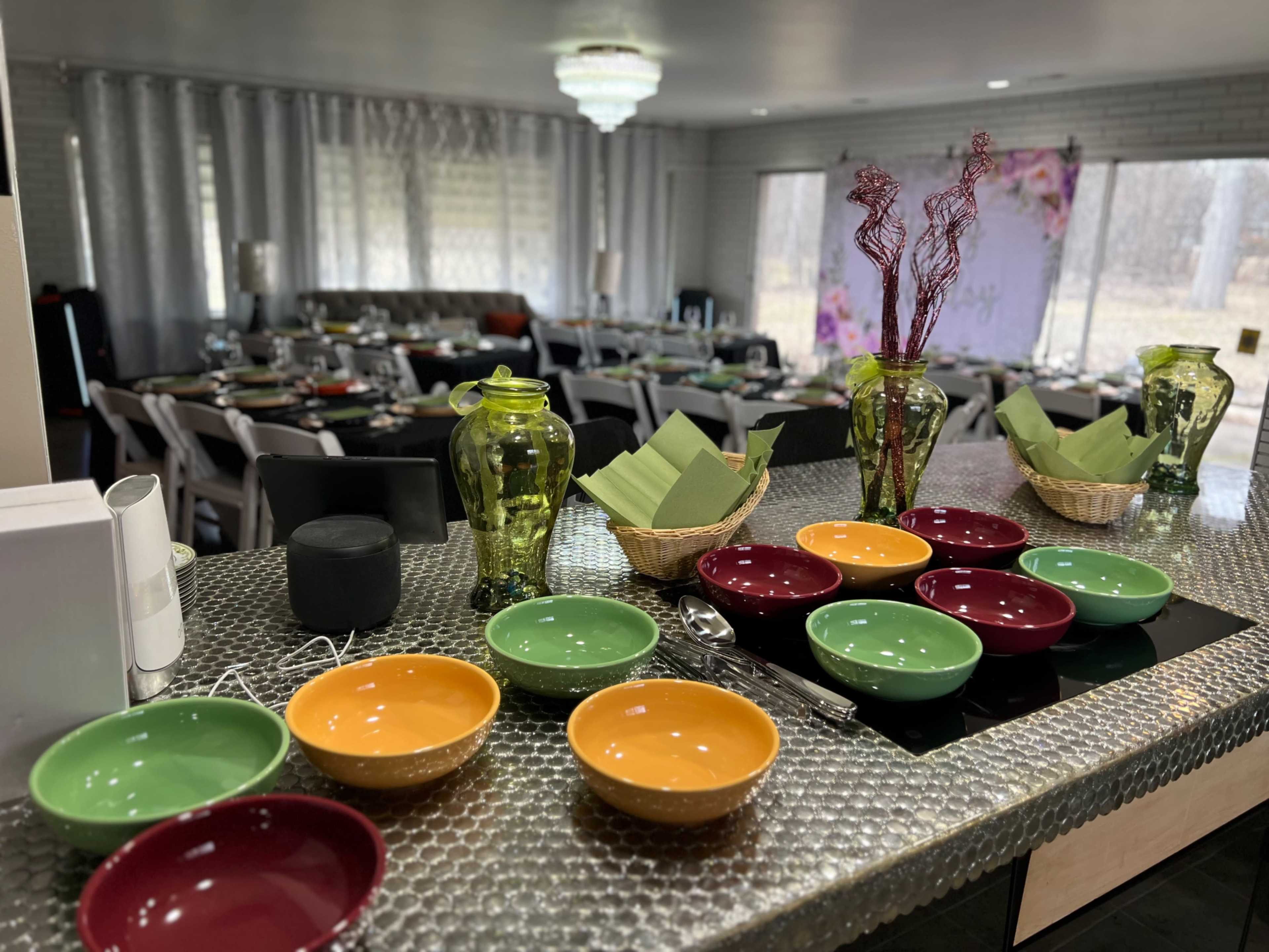 A dining area is set with colorful bowls and decorative items on a countertop, while chairs and tables are arranged for a gathering in the background.