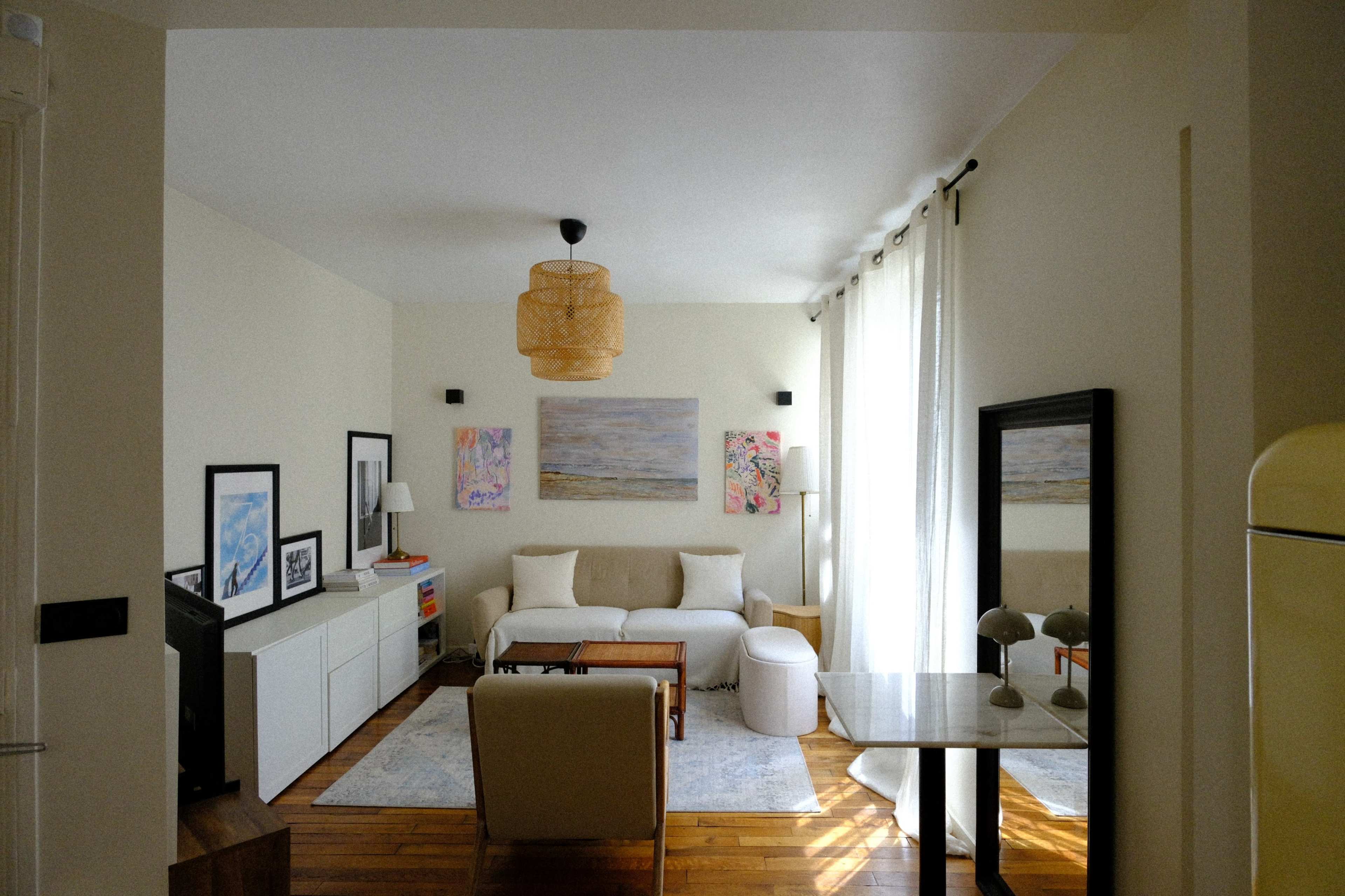 The image shows a bright living room with a sofa, a table, and wall art, featuring large windows with sheer curtains.