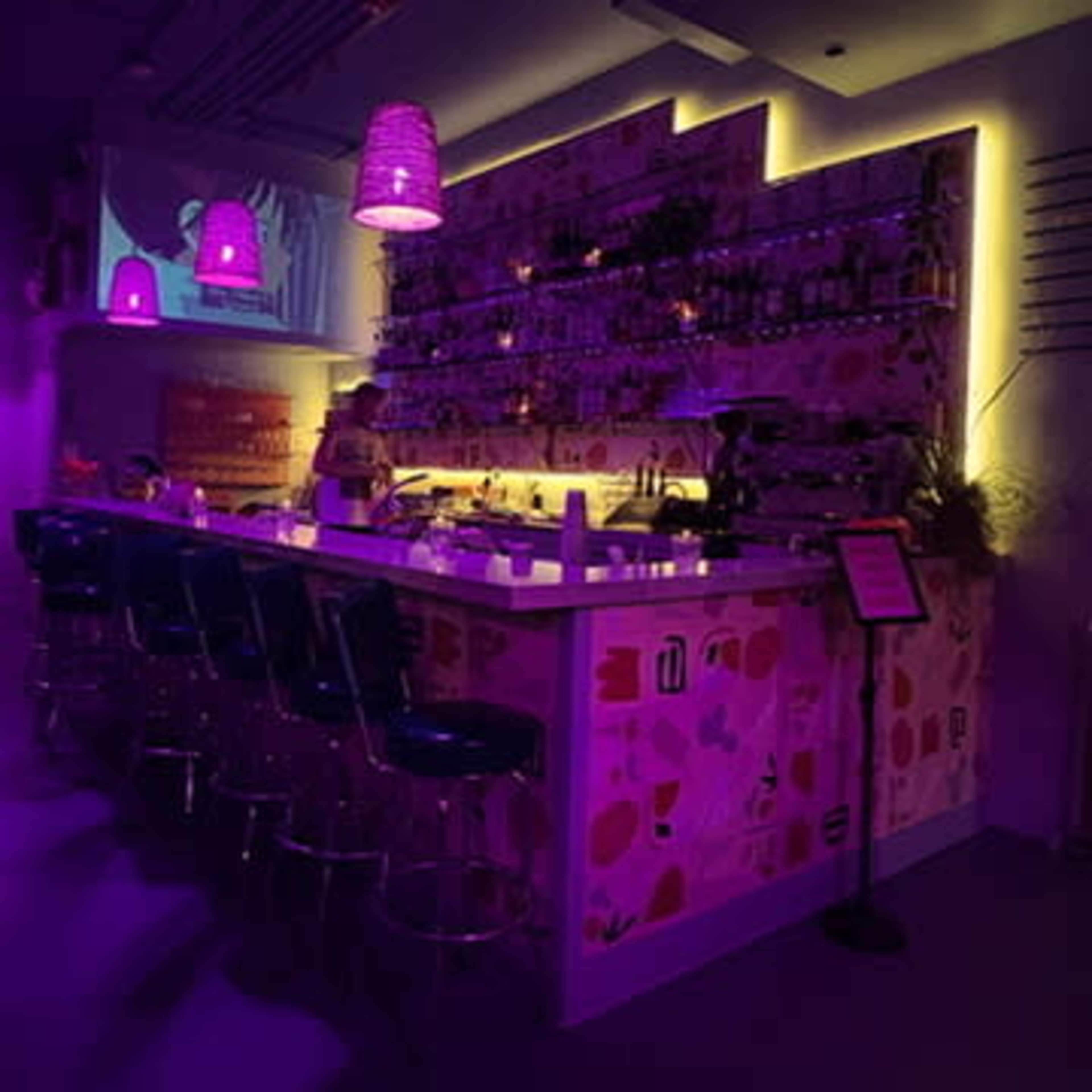 A brightly lit bar features a colorful wall with shelves lined with bottles, illuminated by purple pendant lights.