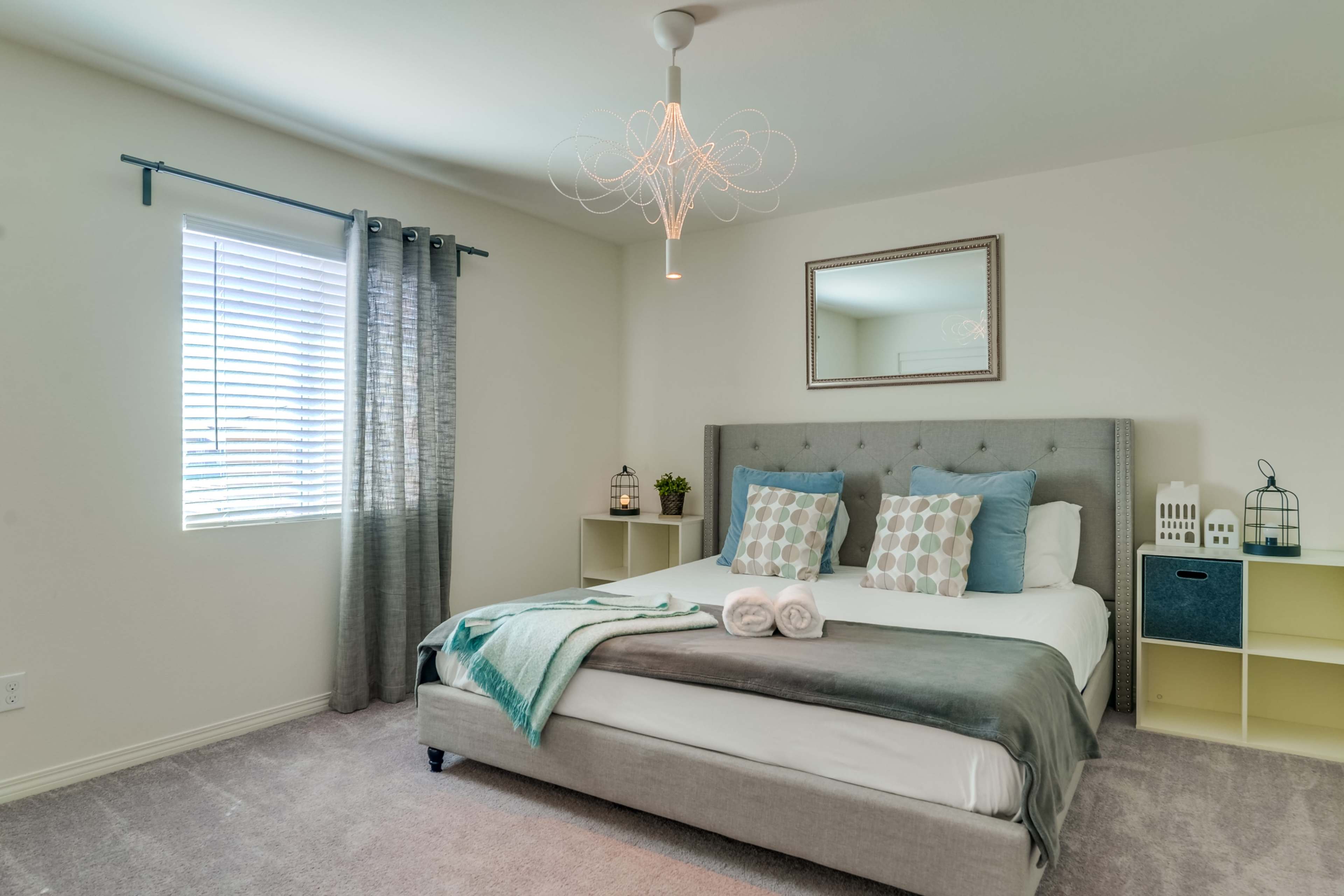 A neatly arranged bedroom features a gray upholstered bed with decorative pillows, a wall mirror, and a window with sheer curtains.