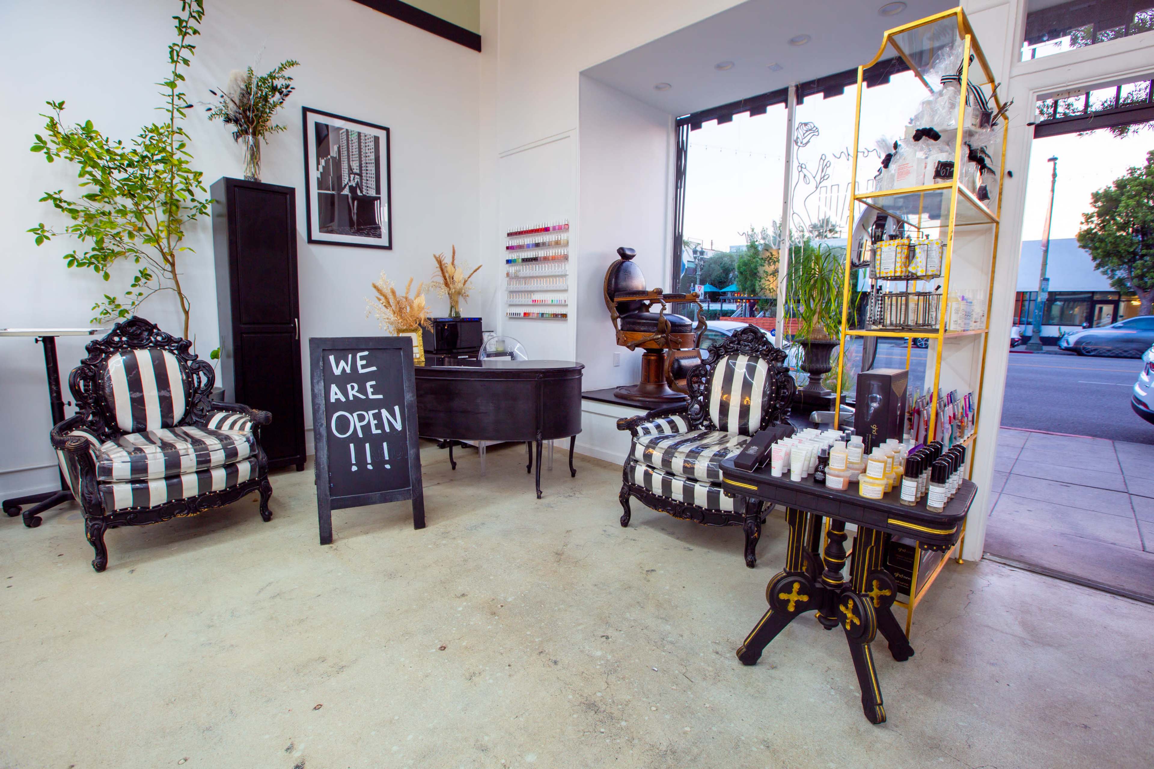 The interior of a shop features two black and white striped chairs, a small black desk, a shelving unit displaying products, and a chalkboard sign that reads "WE ARE OPEN".