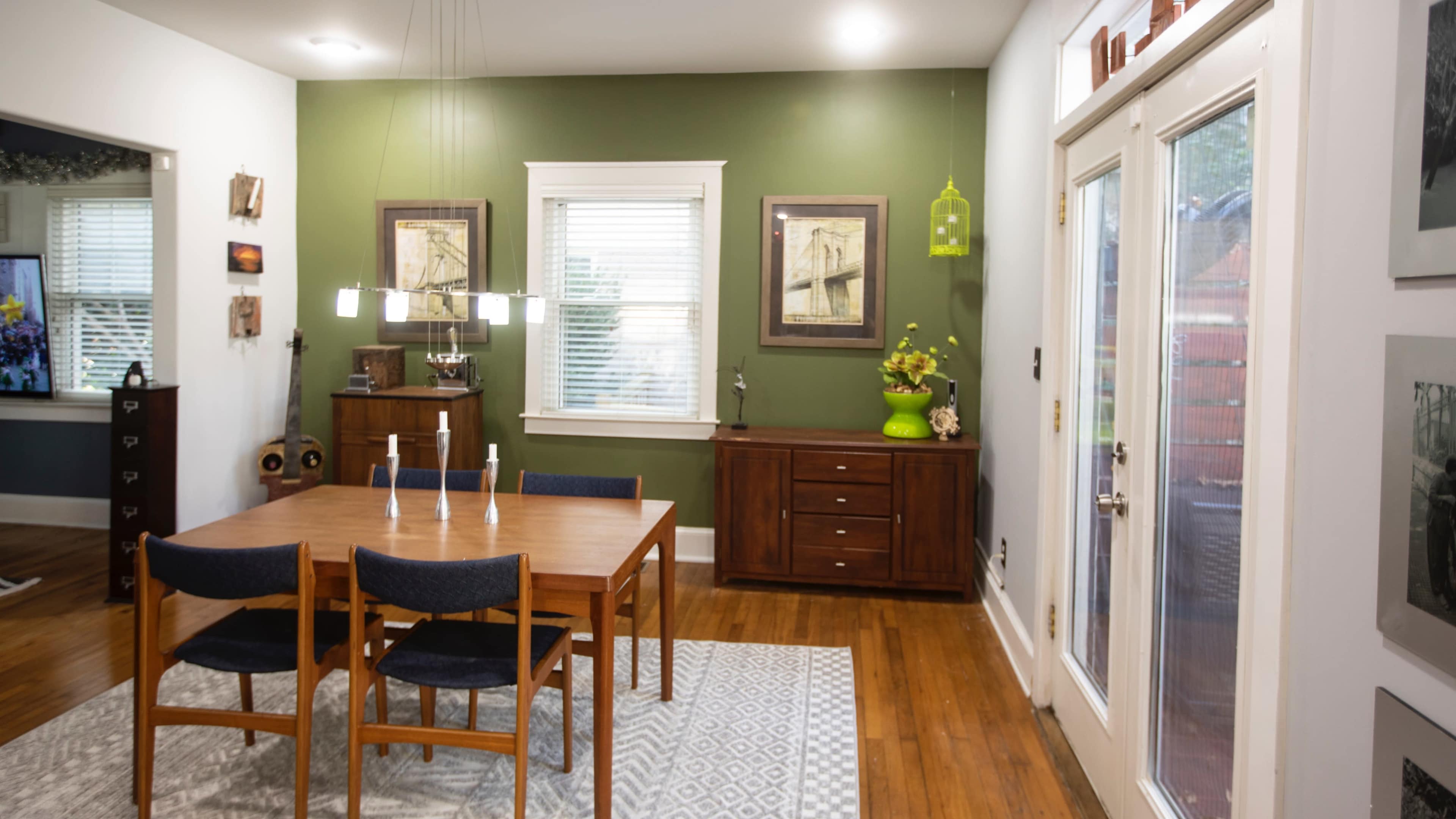 The image shows a dining area with a wooden table and chairs, walls painted in green, and a sideboard against the wall, adorned with framed artworks and a decorative plant.