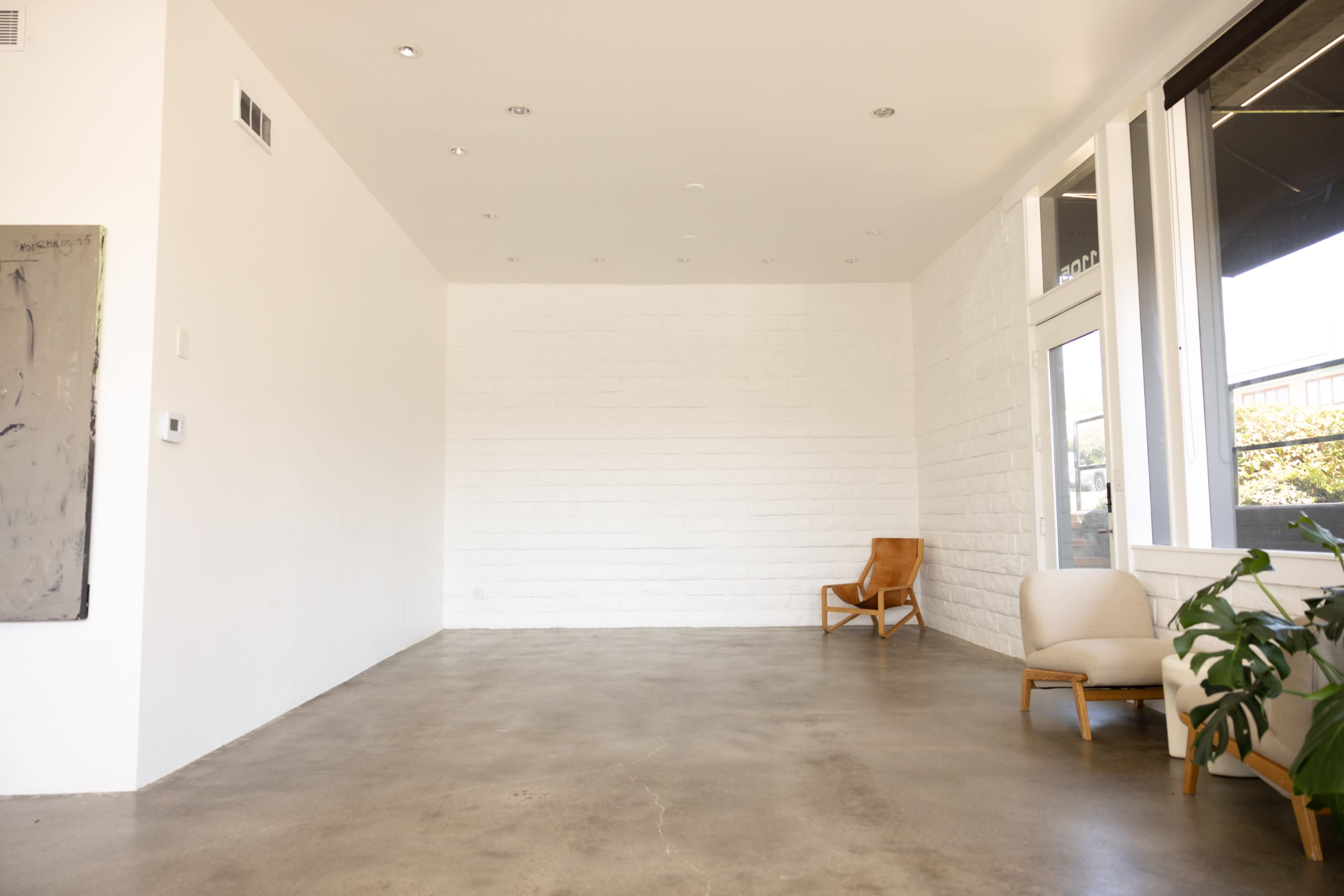 The image shows a spacious, empty room with a concrete floor, a white wall on one side, and two chairs in the corner.