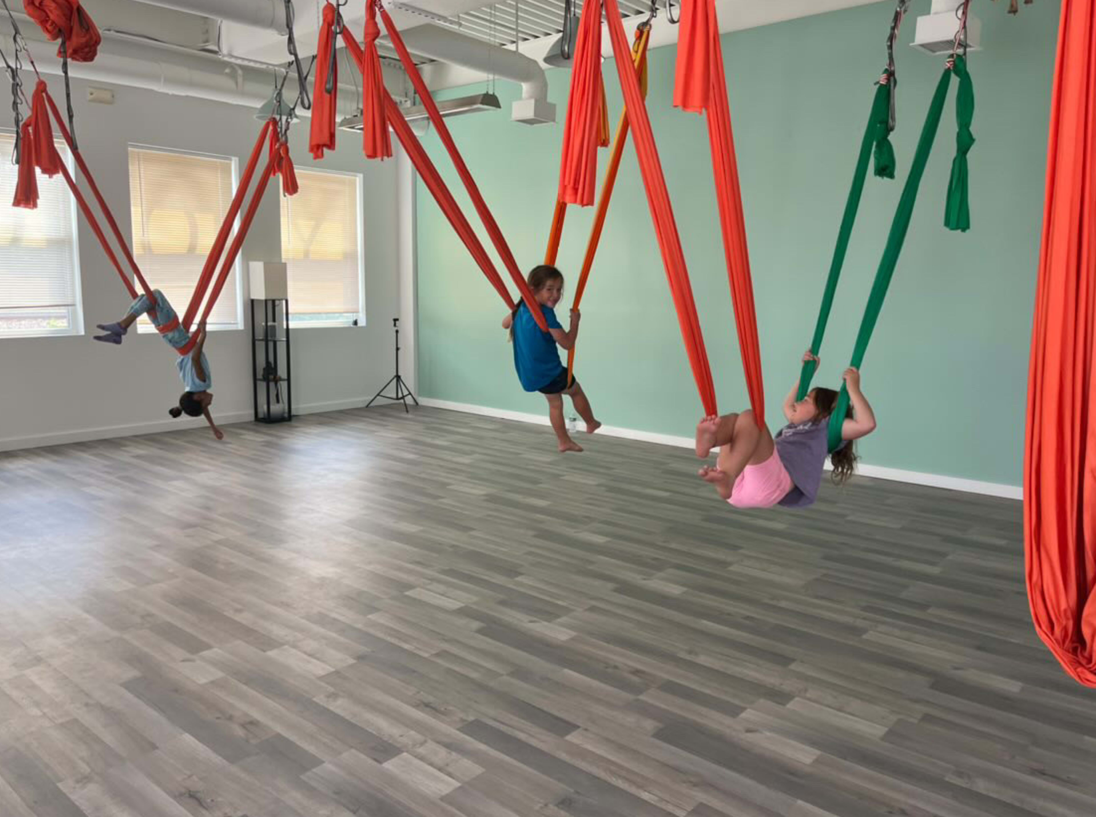 Three children are playing on colorful aerial silk hammocks in a bright, spacious studio.