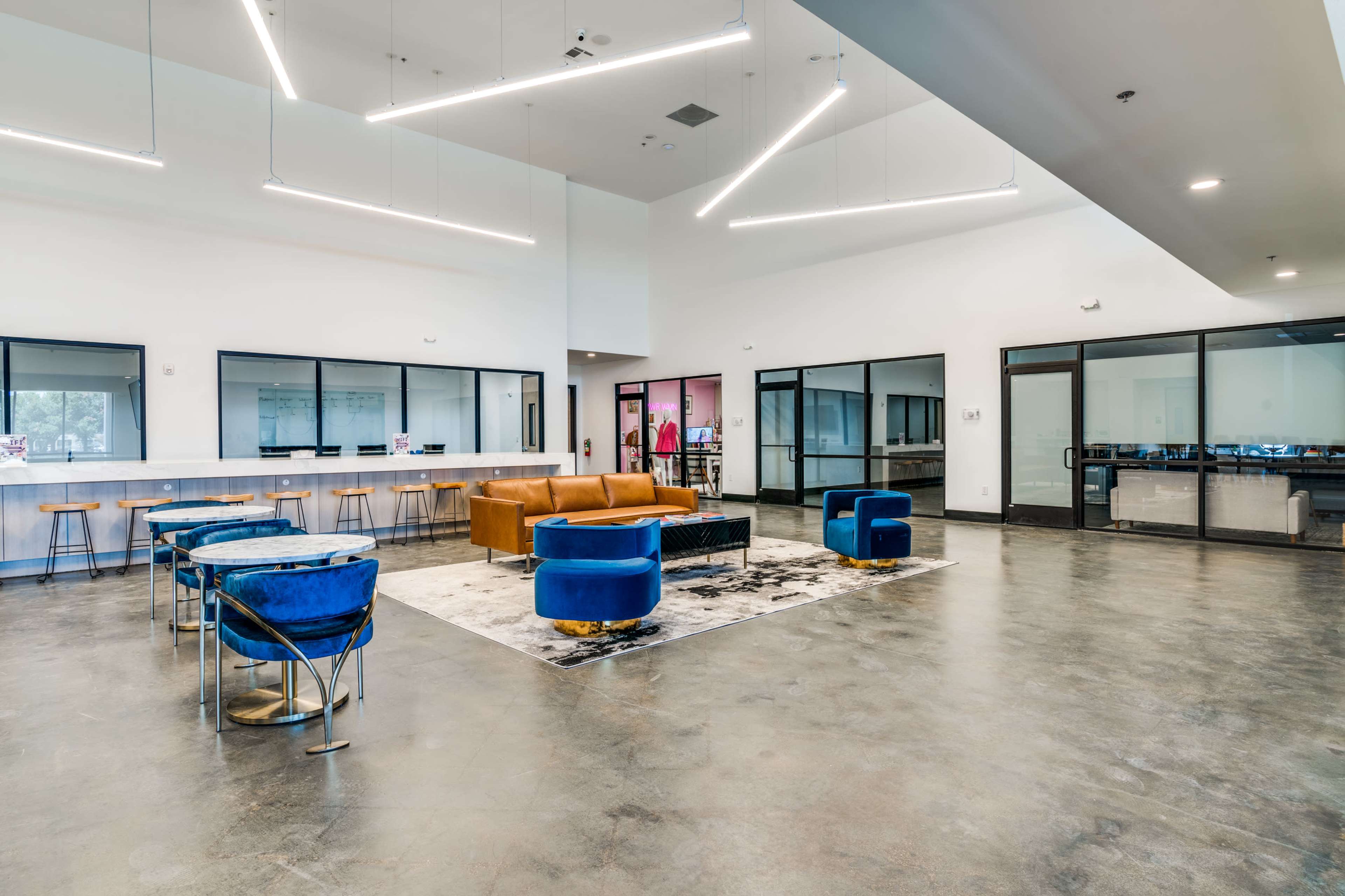 The image shows a spacious interior with a modern design, featuring a brown sofa, blue chairs, a dining area with stools, and large windows.