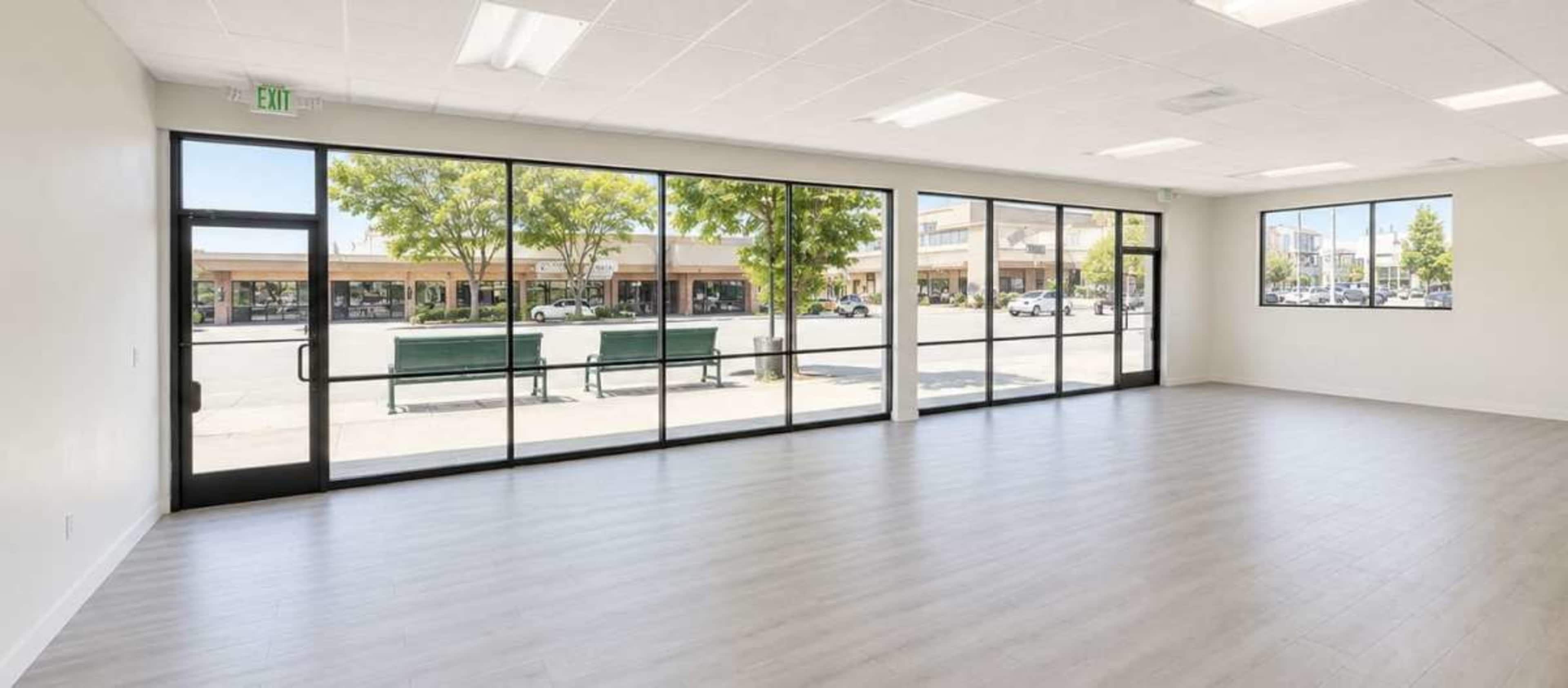 The image shows an empty room with large glass windows overlooking a street lined with parked cars and trees.