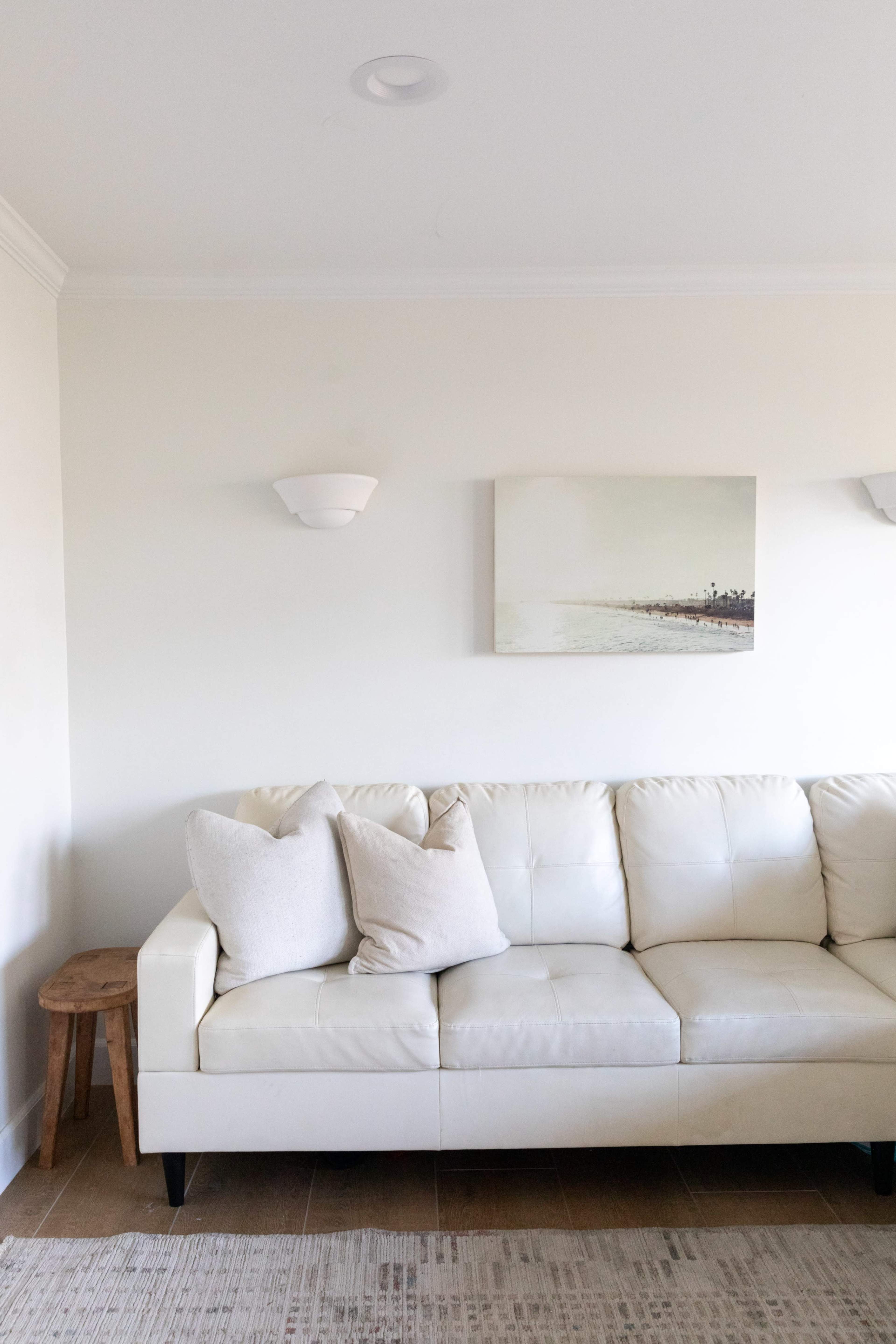 A light-colored sectional sofa with two pillows is placed beside a small wooden stool against a minimalist wall featuring a coastal artwork and sconces.