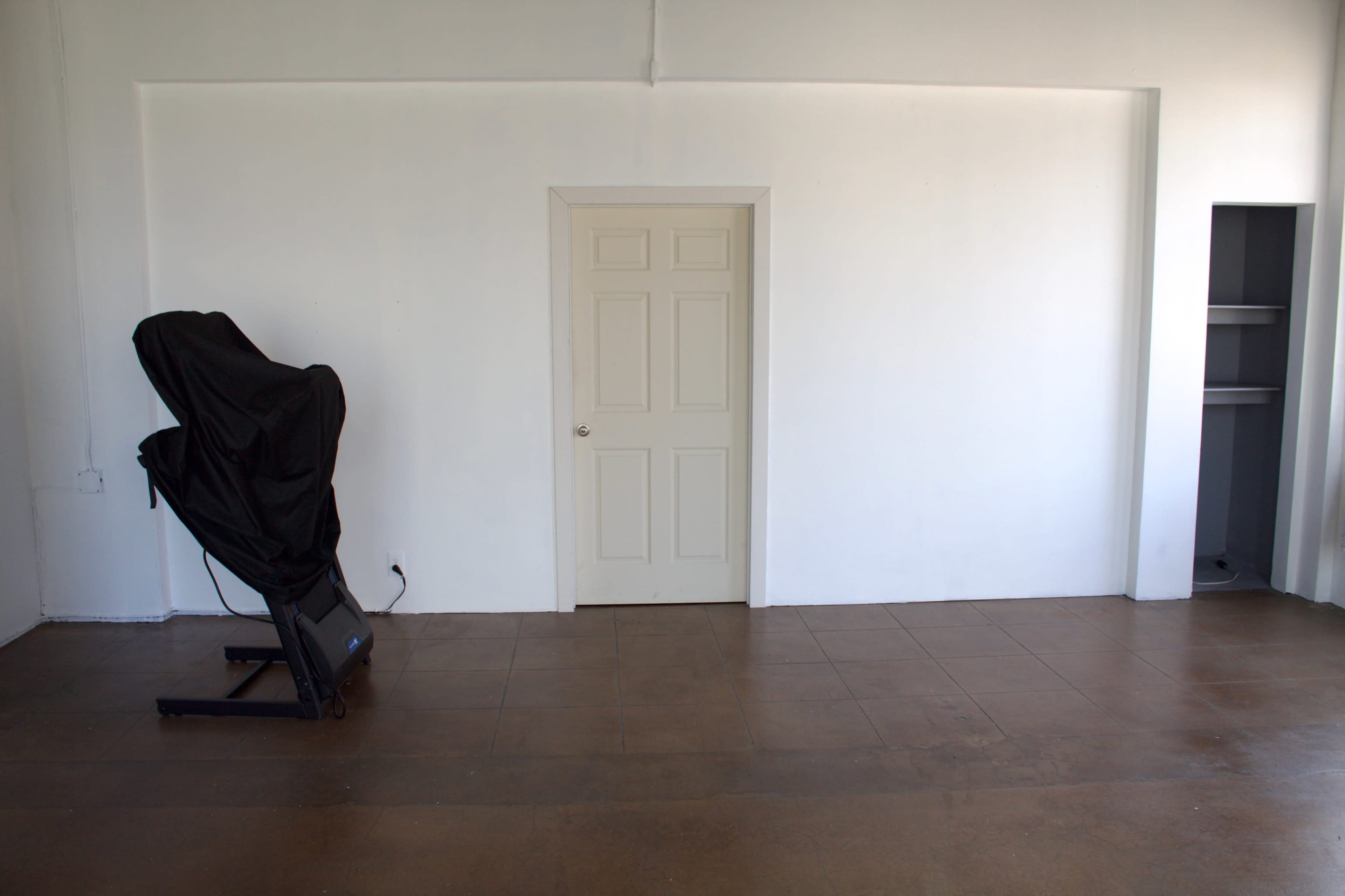 The image shows a minimalist room with a closed door, a partially visible shelf, and a chair covered with a cloth.
