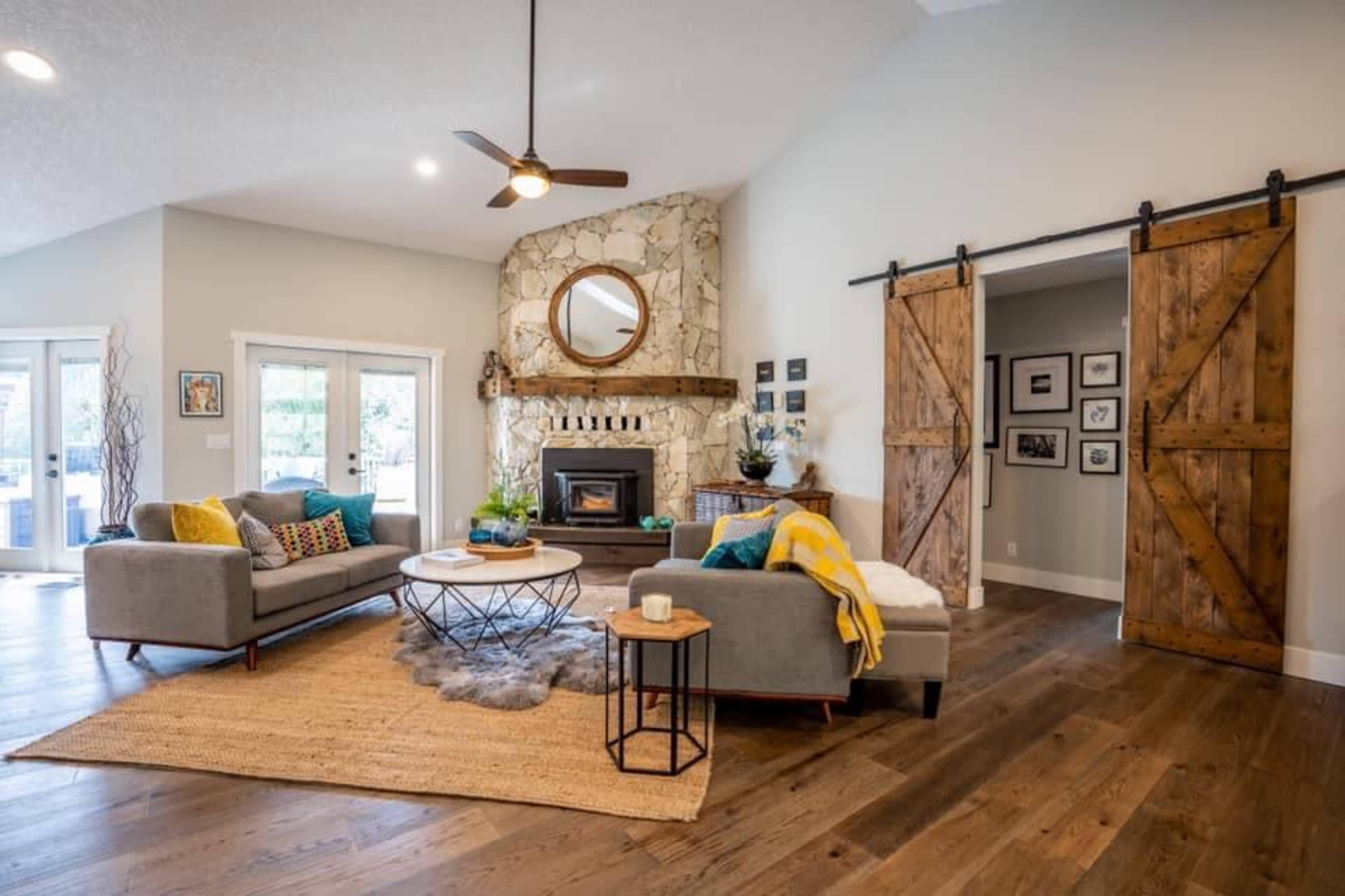 A modern living room with two gray sofas, a circular coffee table, a stone fireplace with a round mirror above it, and sliding barn-style doors, all set on a wooden floor.