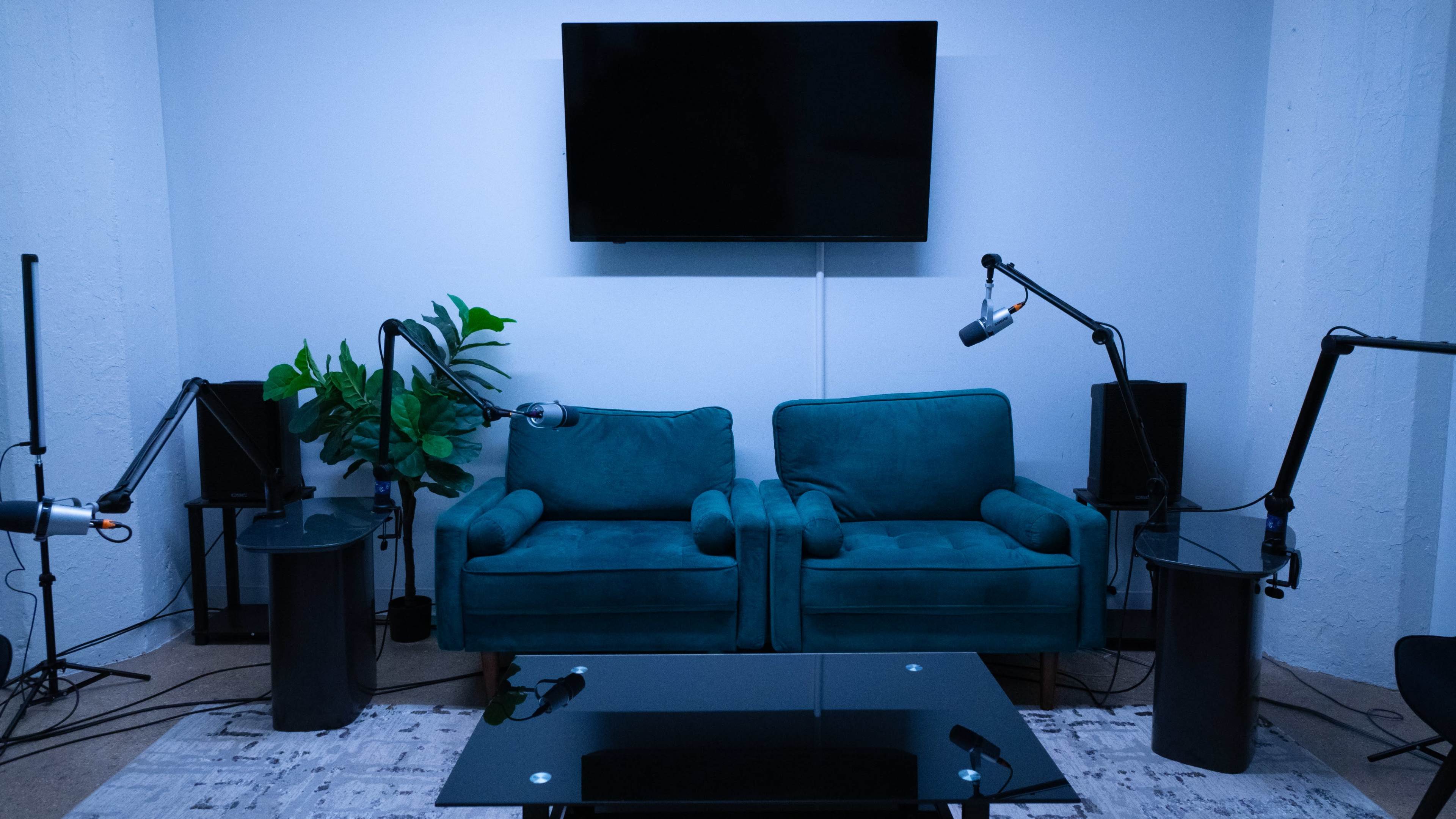 A modern recording studio features a pair of teal couches facing a glass coffee table, surrounded by microphones and a large television on the wall.