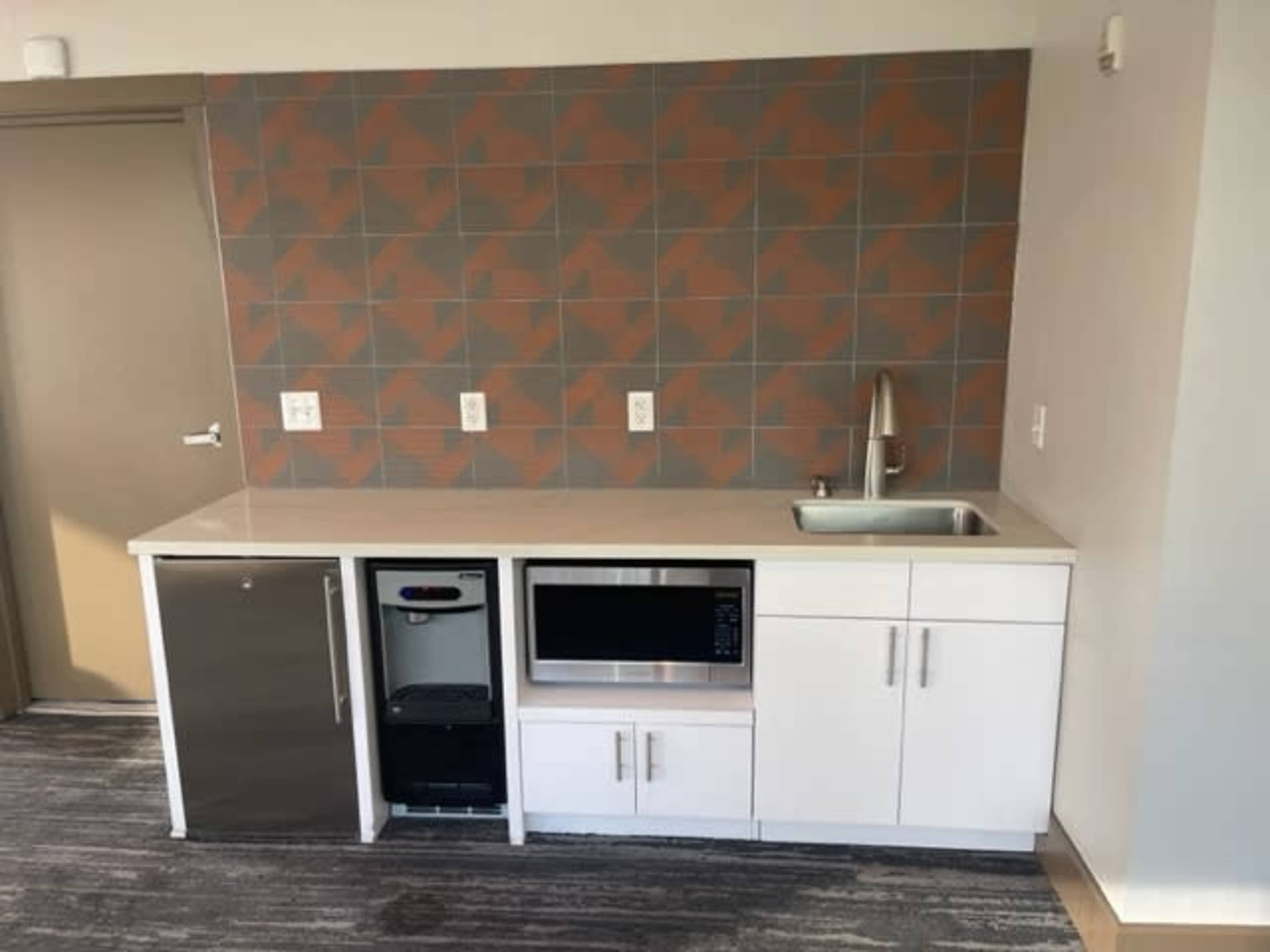 A modern kitchenette featuring a refrigerator, microwave, sink, and cabinets, with a patterned tile backsplash.