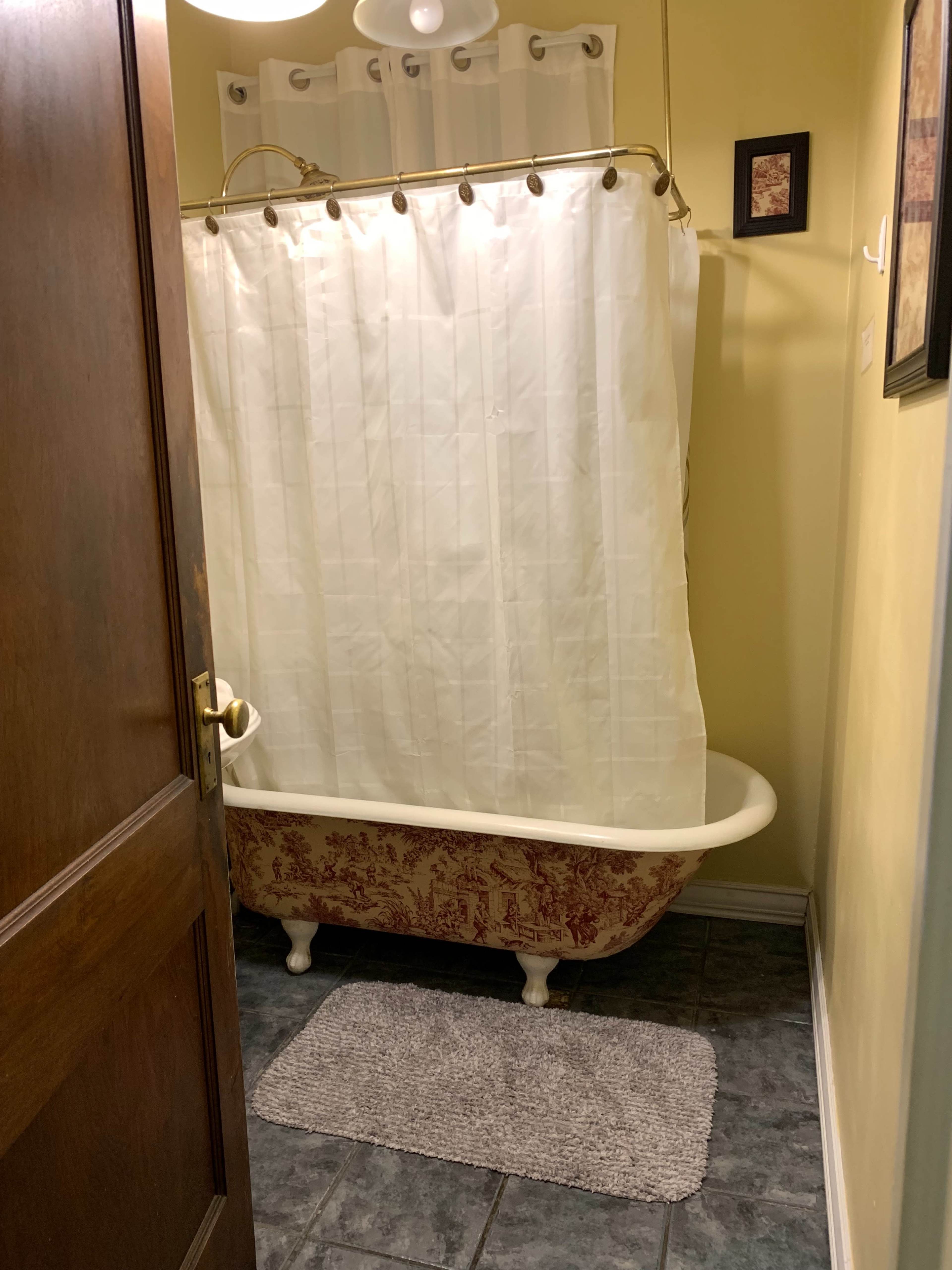 The image shows a bathroom entrance with a vintage bathtub partially obscured by a white shower curtain, set against yellow walls and slate flooring.