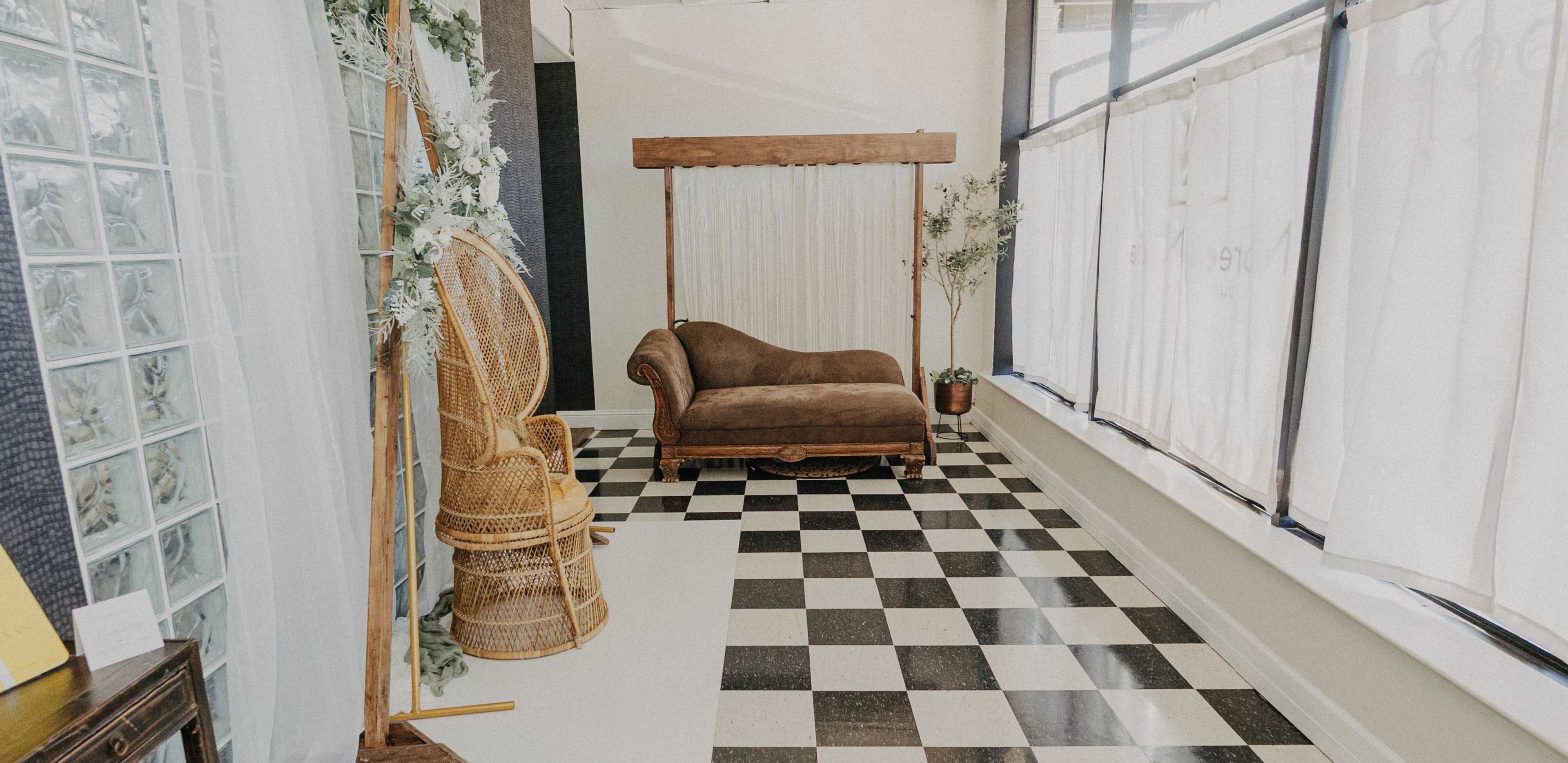 The image shows a room with a checkered floor, a wooden lounge chair draped in fabric, and wicker furniture near large windows covered with sheer curtains.