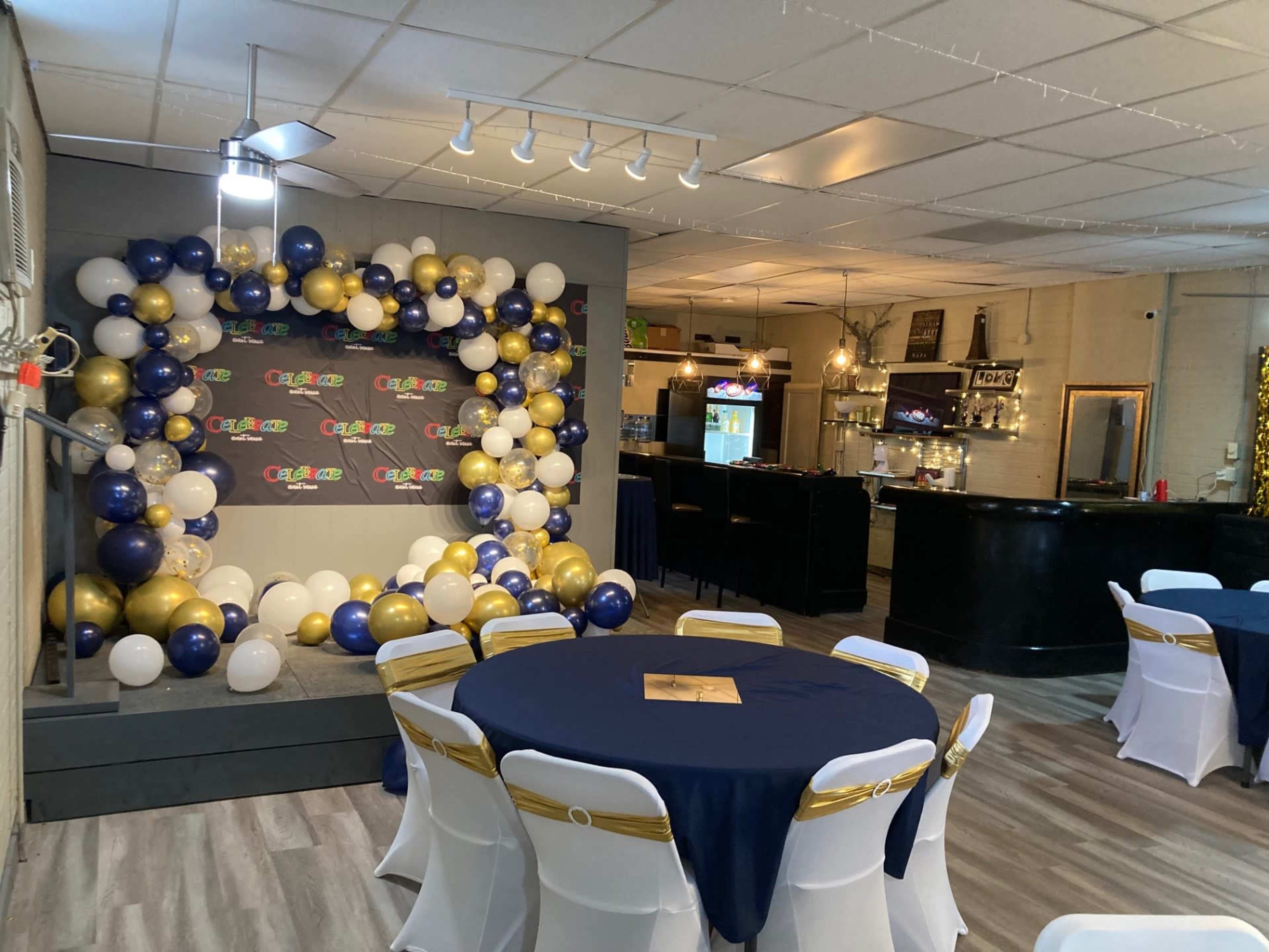 The image shows a decorated event space with tables covered in dark blue cloths and a backdrop of balloons in black, gold, and white.