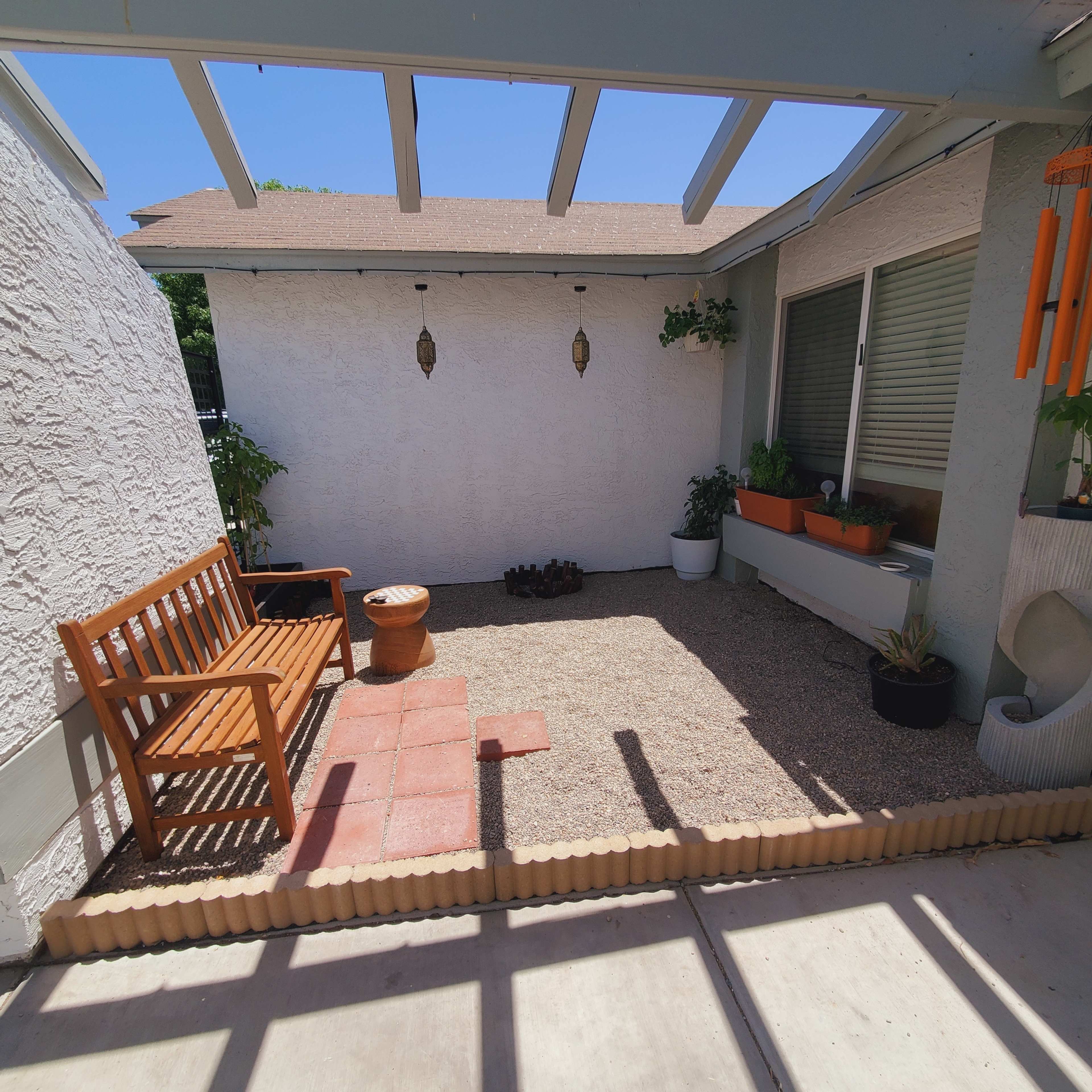 The image shows a small outdoor patio area with a wooden bench, potted plants, and gravel flooring, enclosed by a white wall and a roof supported by beams.