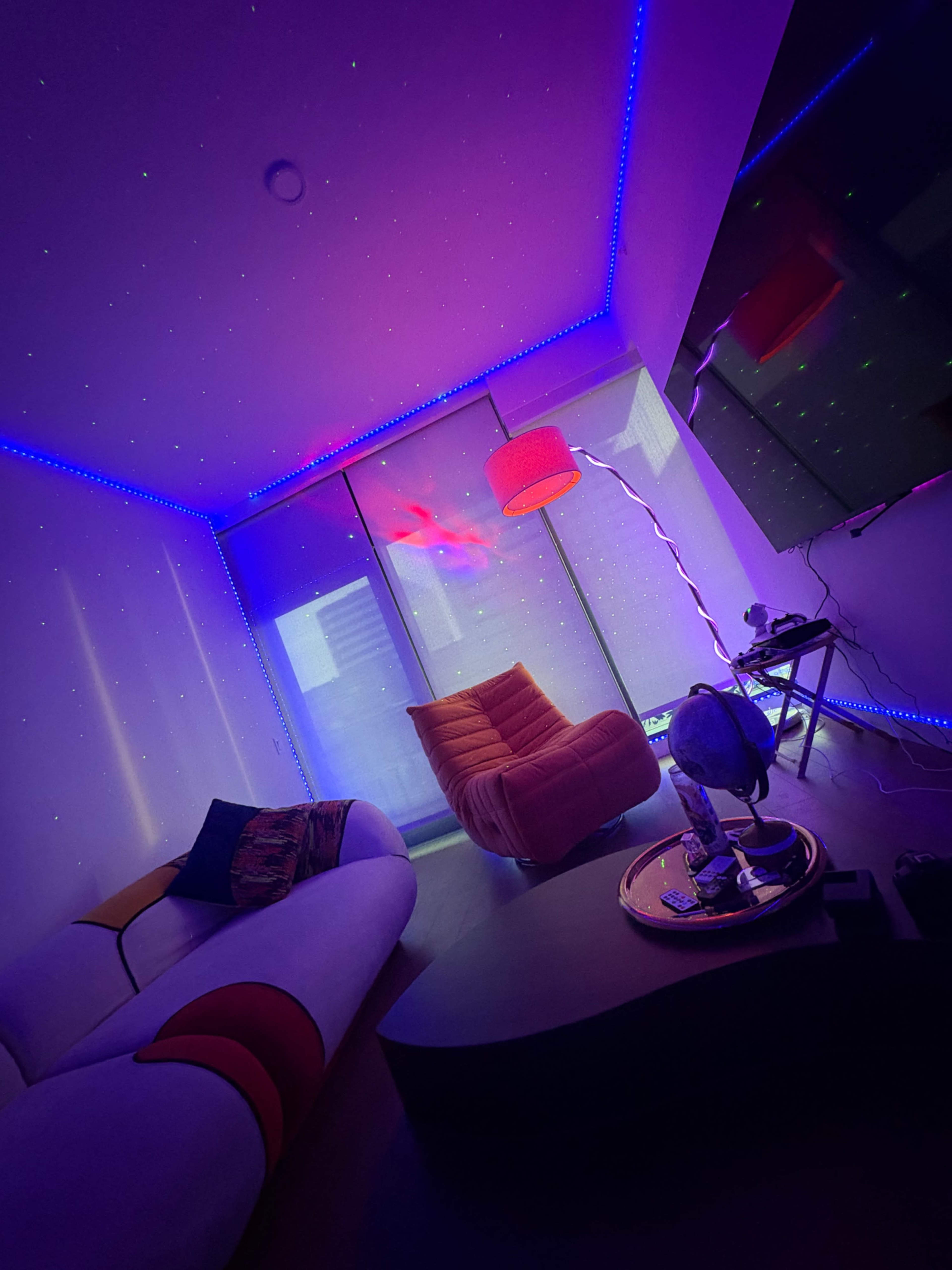 The image shows a modern room featuring colorful LED lighting, a stylish orange chair, and a round coffee table with various items on it.