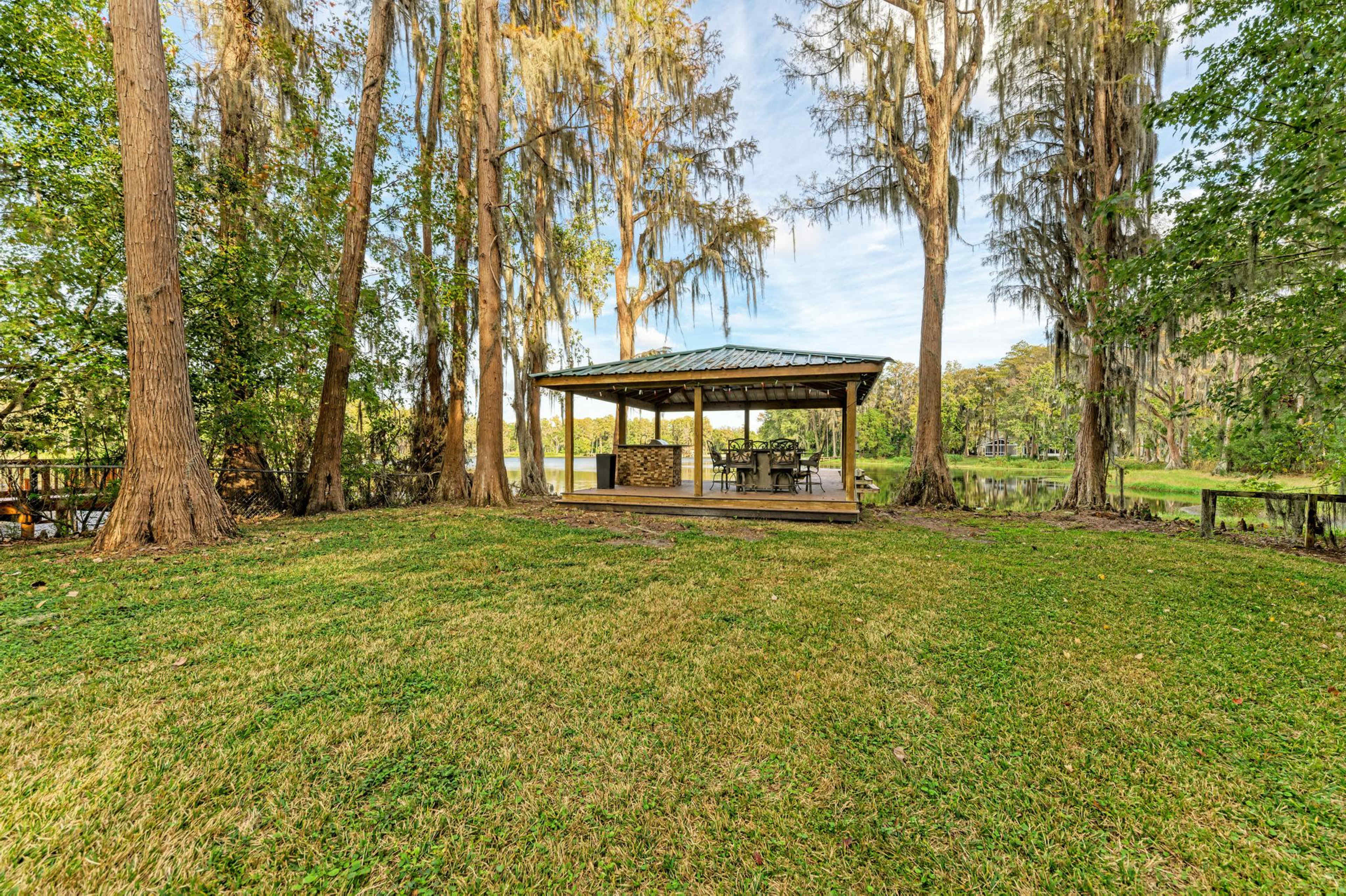 A covered gazebo with a table and chairs is situated on a grassy area near a water body, surrounded by tall trees.