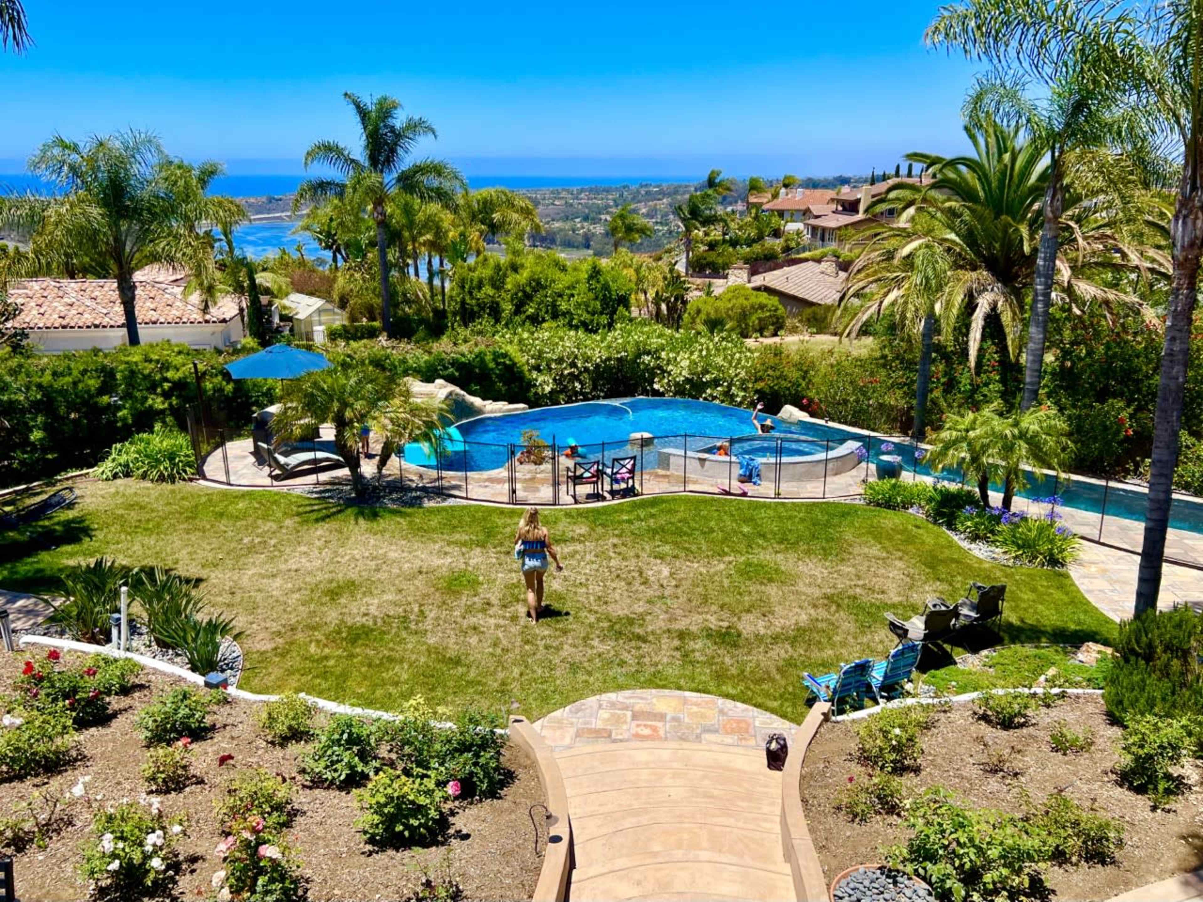 The image shows a backyard with a swimming pool surrounded by palm trees, grassy areas, and a view of the ocean in the background.
